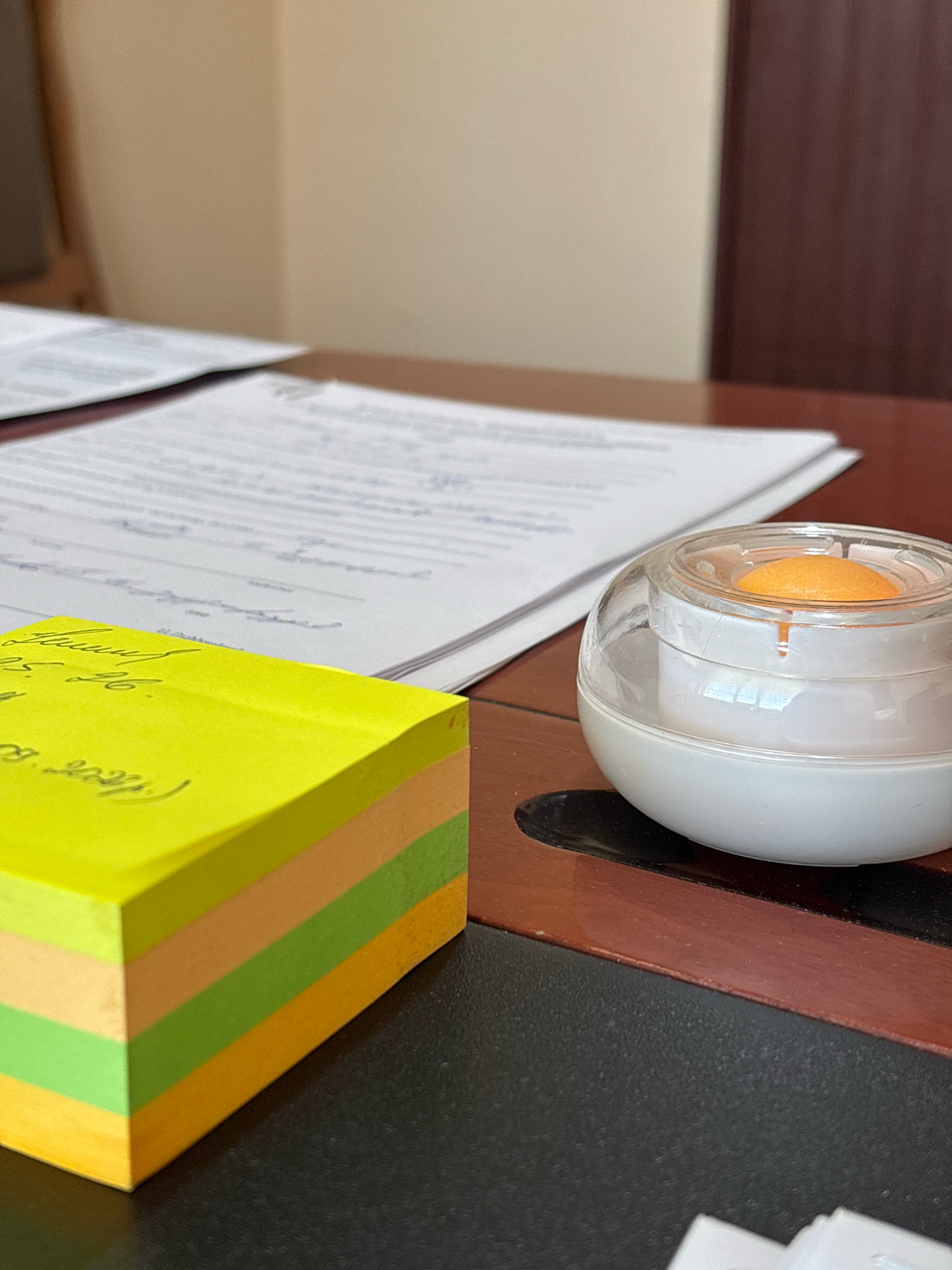 The image depicts a close-up, eye-level shot of a desk in what appears to be an office environment. On the left, a stack of colorful sticky notes, predominantly yellow, is arranged in a corner, with hints of orange, green, and light green visible in the stack. Some handwritten notes are partially visible on the top yellow sticky note. To the right of the sticky notes, a stack of papers with handwritten text is placed on a wooden desk. In the foreground right, a transparent spherical object with a white base contains a small orange ball, resembling a desk toy or dispenser. The background consists of a wall painted in a light beige color, transitioning to a dark brown vertical panel, possibly a door or cabinet. The lighting suggests an indoor setting with diffused natural light. The image does not contain any discernible elements that would indicate its specific location in Ijevan, Armenia.