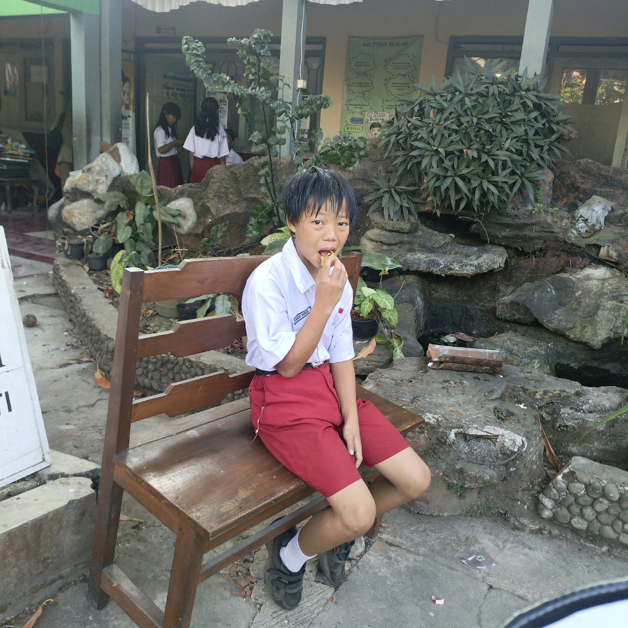 A young boy in a white school uniform shirt and red shorts is sitting on a wooden bench and eating a snack. He is facing the camera with his mouth open as he brings the food to his mouth with his right hand. Behind him, a wall with some plants and a bulletin board is visible. The bulletin board has text in Indonesian, including a poster about bullying. Two other children in school uniforms are visible in the background, walking away from the camera. The setting appears to be an outdoor area of a school in Salatiga, Indonesia, possibly near a decorative rock garden. The bench is made of dark wood with a slatted backrest and seat. The ground is paved with concrete and scattered with fallen leaves and small stones.