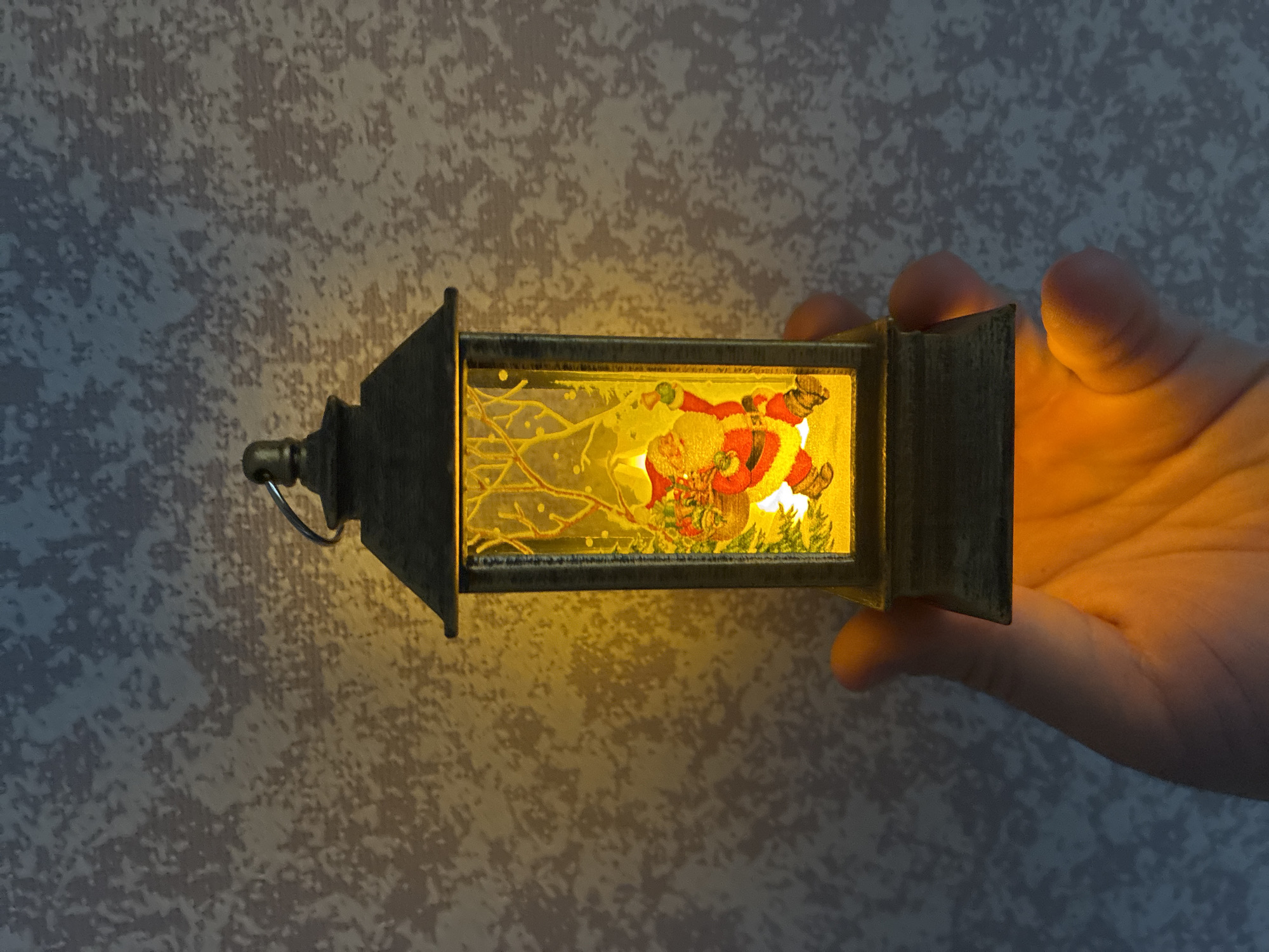 A hand holds an illuminated decorative lantern against a textured wall. The lantern is rectangular with a dark metallic frame and a small loop handle on its top left side. Inside the lantern, a vibrant, backlit miniature scene depicts Santa Claus in his traditional red suit and hat, surrounded by what appears to be snowy branches or a winter foliage design with white and green accents. The entire inner scene glows brightly with a warm yellow light.
This intense illumination from the lantern casts a strong yellow-orange glow on the fingers and palm of the hand holding it on the right side of the frame, highlighting the skin's texture. The light also projects a soft, warm luminescence onto the background wall, which is covered in a light grey wallpaper featuring an irregular, mottled darker grey pattern. The overall impression is an indoor setting, likely in dim ambient light, emphasizing the glowing festive object. No visible text is present in the image.