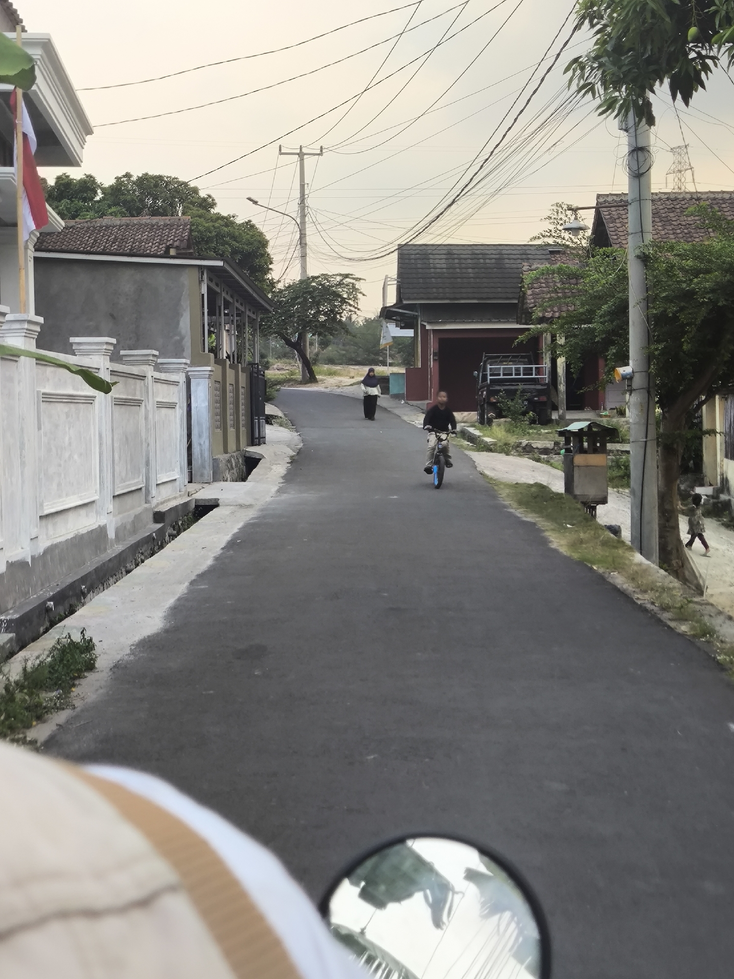The image depicts a street scene in the city of Cilegon, Indonesia, likely during the late afternoon or early evening, indicated by the soft lighting. The perspective is from a vehicle, with a rearview mirror and part of the vehicle's interior visible in the foreground. The street is paved and slopes slightly upwards.
On the left side of the street, there is a white decorative fence with pillars and recessed panels, followed by a building with a tiled roof and a covered walkway supported by columns. A red and white flag, possibly the Indonesian flag, is mounted on a pole on the corner of a building.
On the right side of the street, there are houses with tiled roofs and trees. A utility pole with numerous electrical wires is prominent. A garbage bin with a green roof is situated on the sidewalk.
Several people are present in the scene. A person is riding a bicycle with blue wheels down the street, wearing dark clothing. Further up the street, another person in a hijab is walking. A smaller child is visible further down the right side of the street, walking on the sidewalk. A black pickup truck is parked in front of one of the houses. The environment appears to be a residential area.