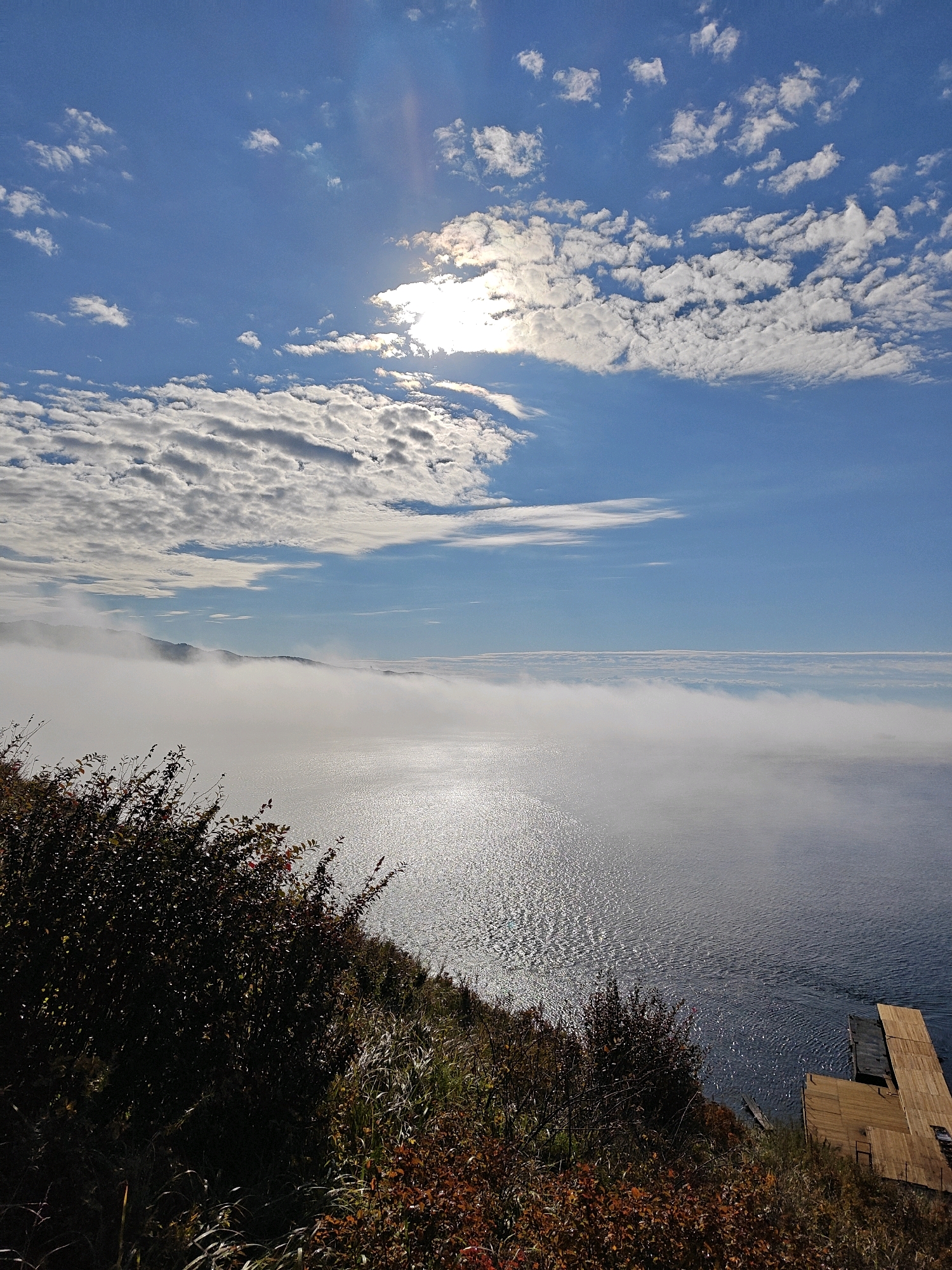The image depicts a vast body of water, likely Lake Baikal in Russia, under a bright, partially cloudy sky. The sun is visible, partially obscured by clouds, casting a shimmering reflection on the water's surface. A thick layer of fog or mist hovers over the water in the middle ground, obscuring a distant shoreline with rolling hills. In the foreground, a dense, dark green bush with some reddish leaves grows on a slope overlooking the water. Sparse, dry grass is also visible on the slope. To the right, a wooden dock structure extends into the water. The scene is calm and natural, with no people or animals present.