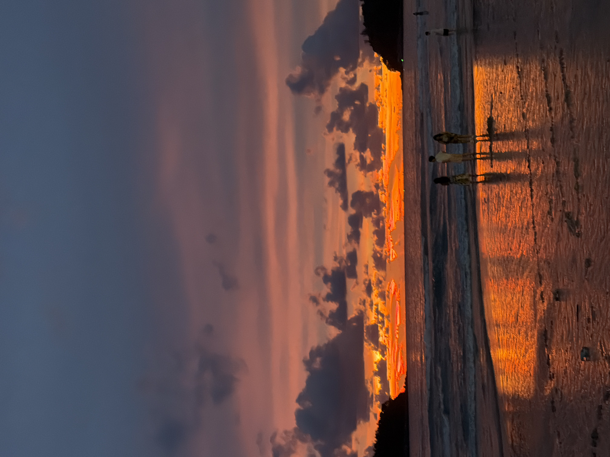 A wide shot captures a breathtaking sunset at Karon Beach in Karon, Thailand. The sky is a vibrant canvas of deep purples, fiery oranges, and soft pinks, with dramatic dark clouds scattered throughout. The sun is low on the horizon, casting a brilliant orange glow across the ocean and the wet sand. Gentle waves lap at the shore, reflecting the colorful sky.
In the foreground, three individuals stand in the shallow water, their silhouettes distinct against the illuminated sea. To the right, a lone figure stands further out in the water, and another person is visible in the distance on the beach. Dark, tree-covered headlands frame the left and right sides of the scene, adding depth and context to the beach setting. The wet sand in the foreground is a mirror, doubling the intensity of the sunset's reflection.