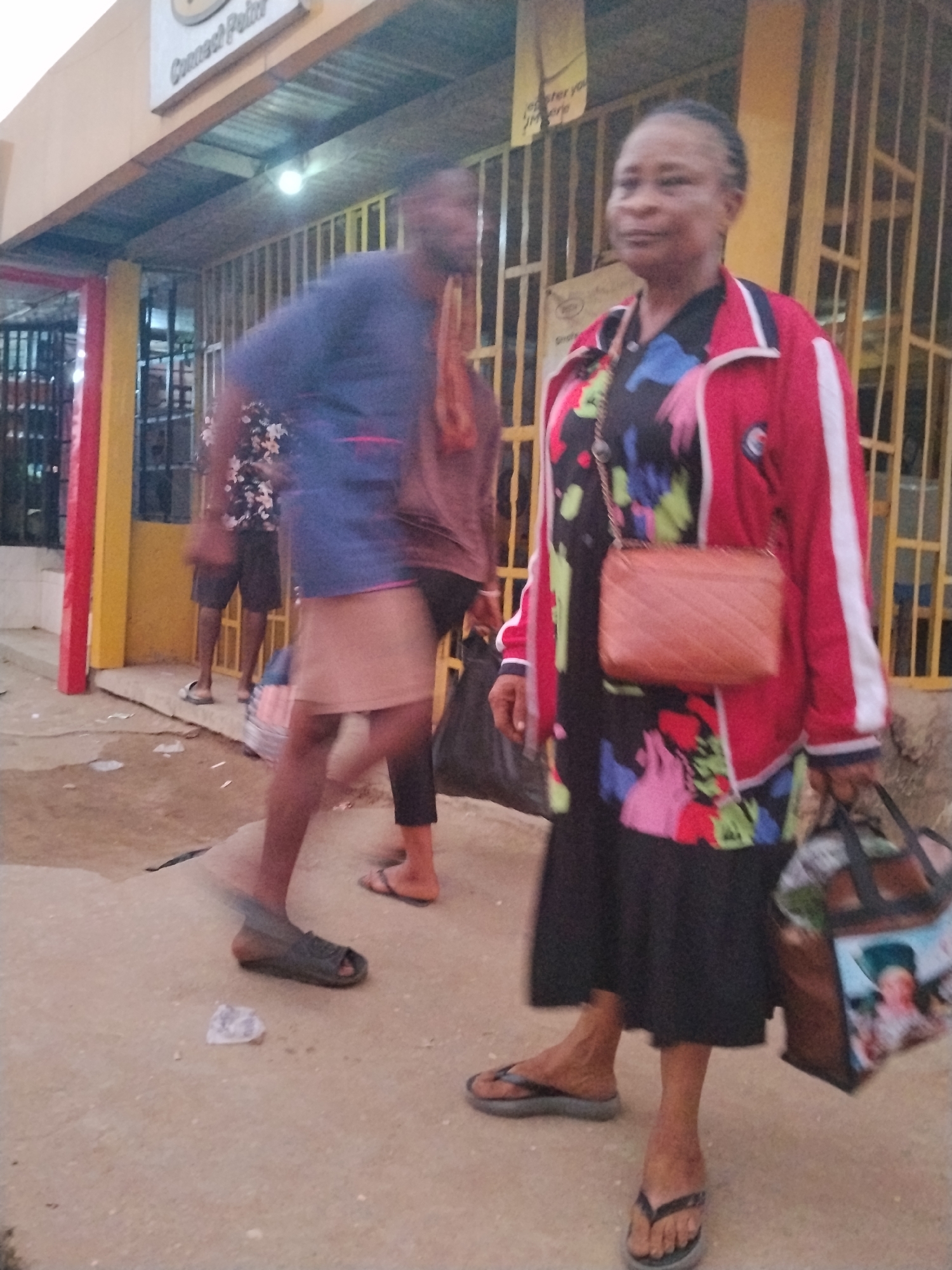 The image captures a street scene in Ura Nta Umuarandu, Nigeria. An older woman stands in the foreground, wearing a red and white jacket over a floral dress, a brown purse, and carrying a large bag. Several other people are in the background. A man in a blue shirt walks past. The scene takes place in front of a building with a sign that reads "Connect Point". The ground is concrete. The overall impression is one of a busy, everyday street scene.
