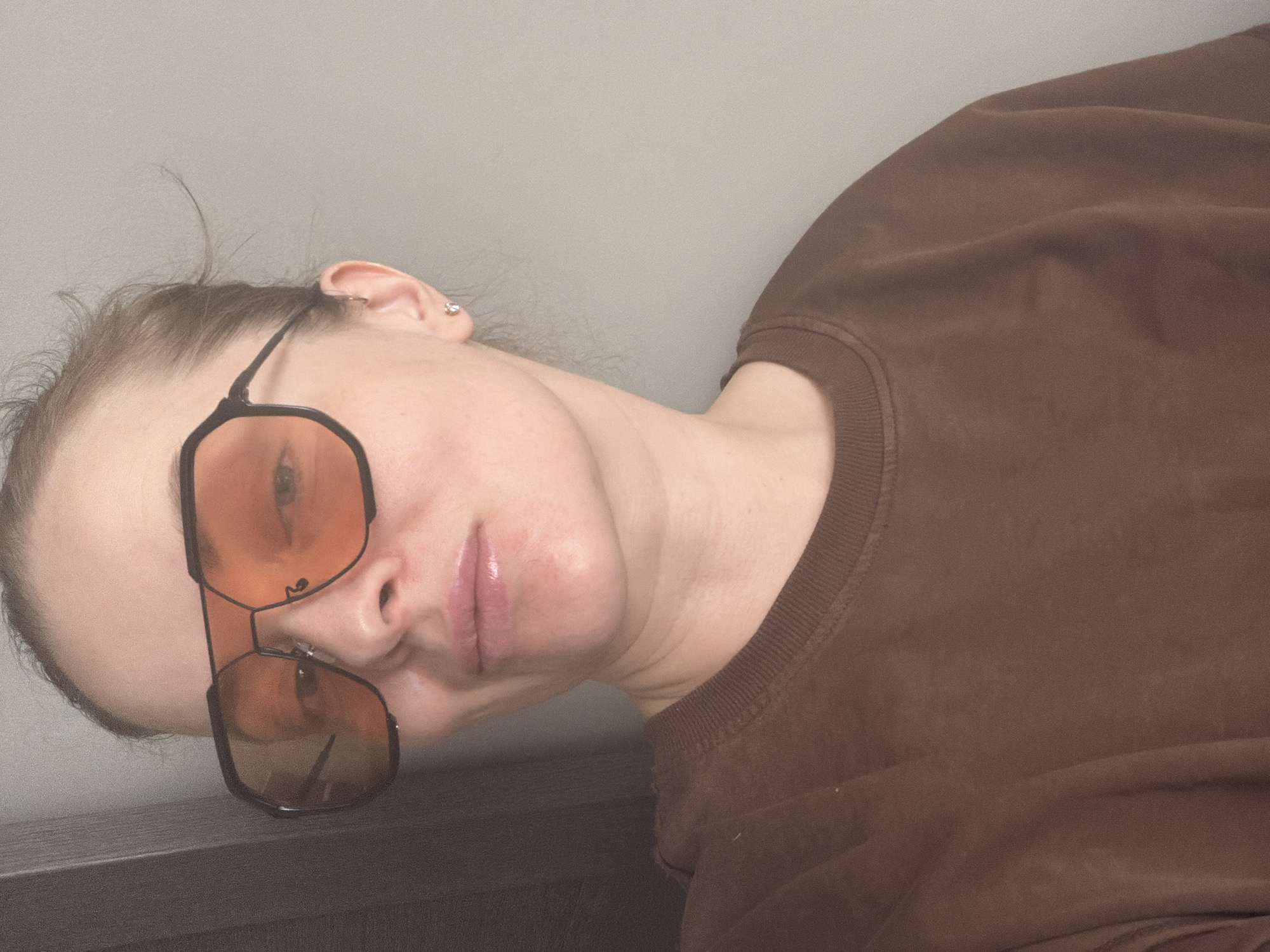 A woman with fair skin and light brown hair, wearing a brown sweatshirt and black-framed sunglasses with orange lenses, is looking toward the camera. She appears to be lying on her side. The background is a neutral, pale grey wall. The edge of what might be a dark-colored piece of furniture is visible in the lower left corner of the image. There's no indication of the location being Moscow, Russia, in the scene.
