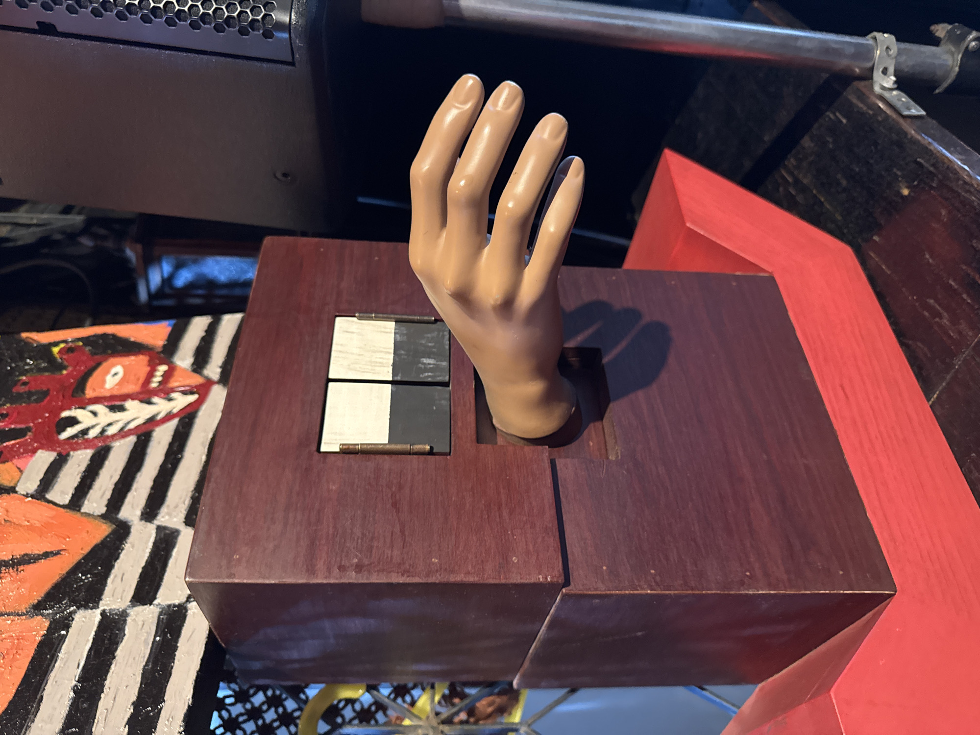 An artificial hand emerges from a box with black and white shutters. The box is dark brown. A red frame is visible next to the box. The background is cluttered with mechanical and wooden parts. The location is Bangkok, Thailand.