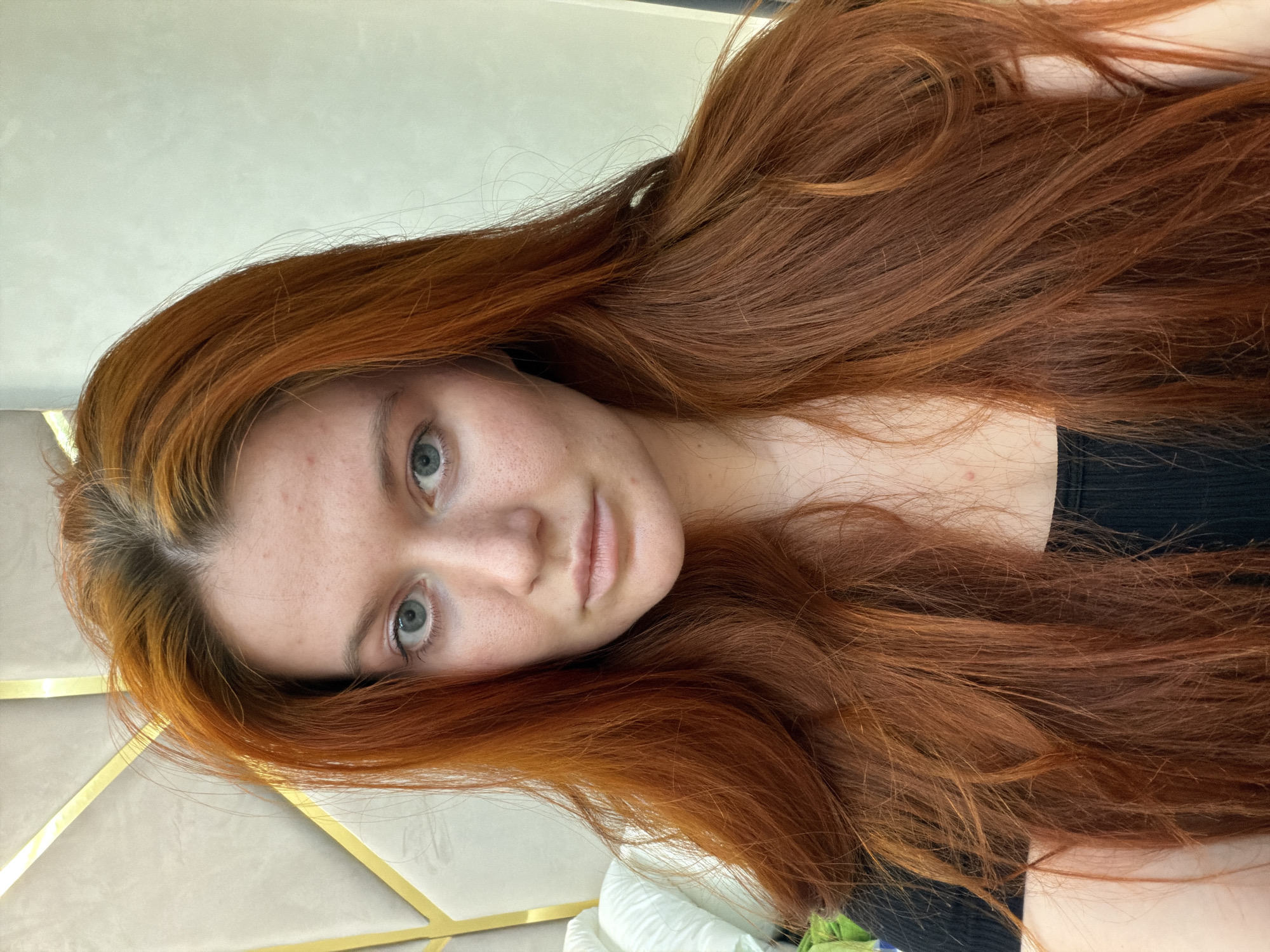A young woman with long, bright red hair, fair skin with freckles and some blemishes, and light blue eyes is shown in a close-up portrait. She is looking directly at the camera with a neutral expression. Her hair is styled down and appears somewhat tousled. She is wearing a black top. The background consists of a neutral-colored wall with geometric gold accents, and a hint of white bedding on the left side. The setting appears to be indoors, possibly a bedroom or a well-decorated living space. There is no information in the image to indicate it is located in Krasnodar, Russia.