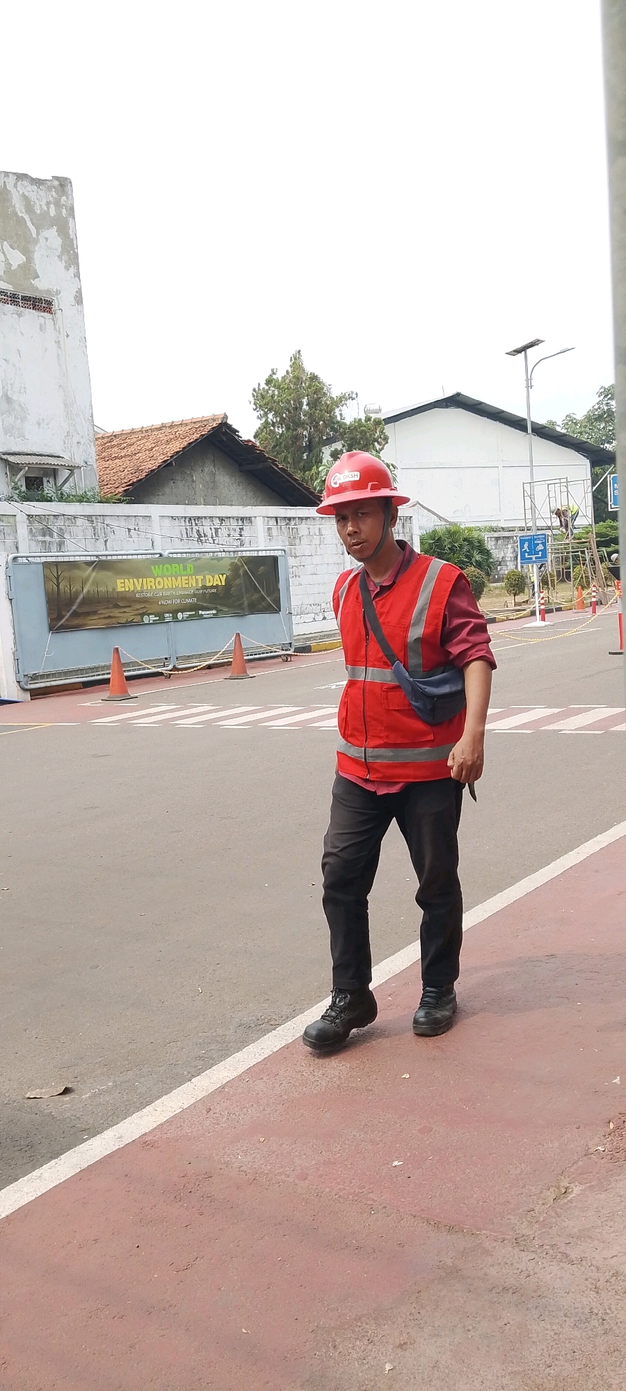A man wearing a red hard hat and a red reflective vest over a maroon shirt and black pants walks on a sidewalk adjacent to a road. He is also wearing black work boots and a dark blue sling bag across his chest. In the background, a banner is displayed on a gate that reads "WORLD ENVIRONMENT DAY RESTORE OUR EARTH. ENSURE OUR FUTURE. #NOW FOR CLIMATE." The scene appears to be outdoors in a somewhat industrial or commercial area. There are orange traffic cones visible near the gate, and a white and red striped pedestrian crossing is marked on the road. A white building with a tiled roof is visible behind the gate, as are some trees and a lamppost. The overall setting suggests an urban environment.