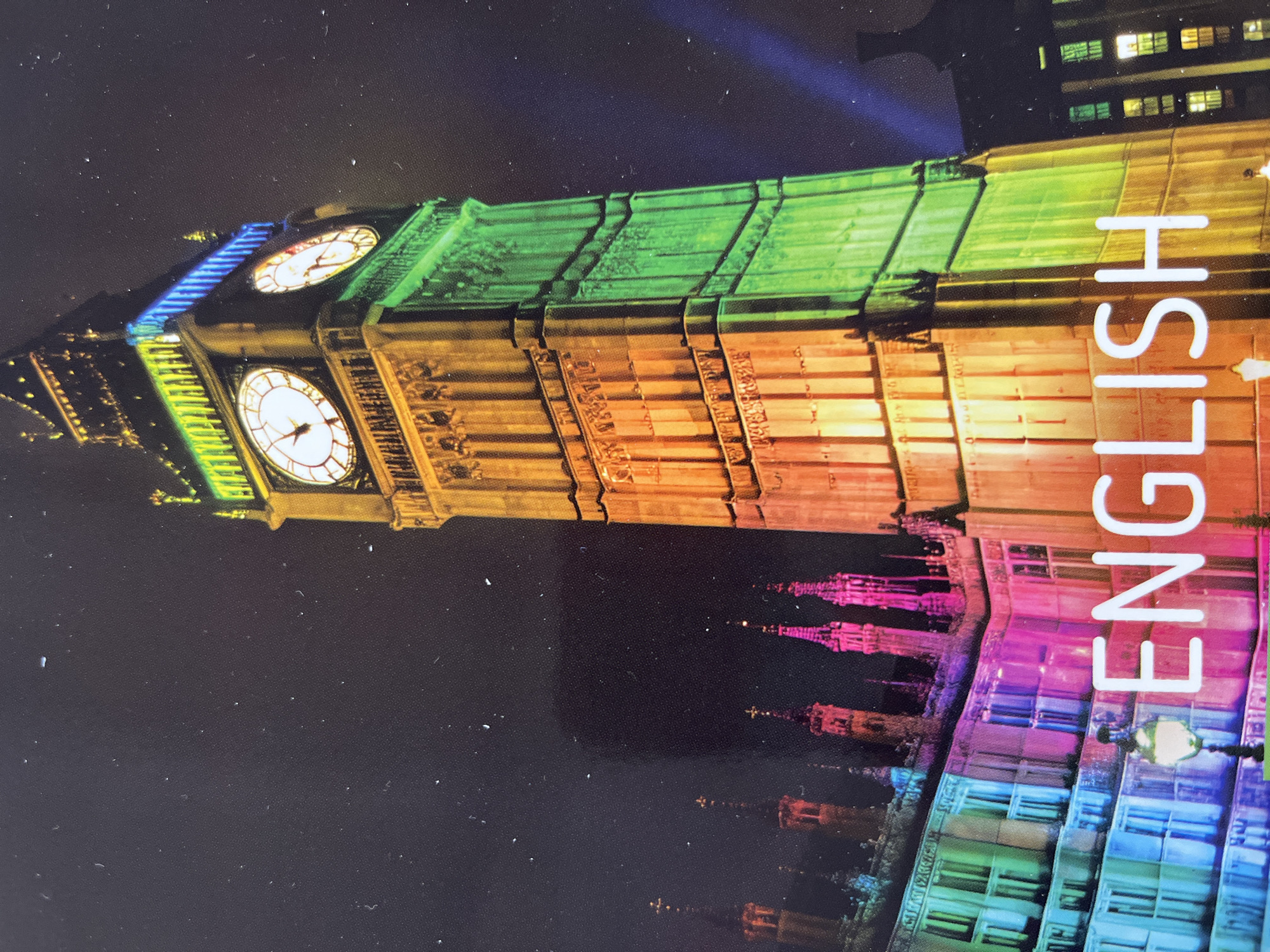 The image shows the Elizabeth Tower (commonly known as Big Ben) in London, UK, illuminated at night with colorful lights. The tower is lit with a gradient of colors, from orange and yellow on the left to green and blue on the right. The Palace of Westminster is visible to the left of the tower, also illuminated with vibrant colors. White text that reads "ENGLISH" is prominently displayed in the foreground, superimposed over the colorful lights of the buildings. The sky is dark and speckled with what appear to be small white specks, possibly dust or imperfections on the image itself, or distant stars. There are no people or discernible actions depicted in the image. The scene is an exterior nighttime view of a famous landmark. The prompt's request to include the city of Kharkiv, Ukraine, is factually incorrect as the image clearly depicts Big Ben in London, UK.