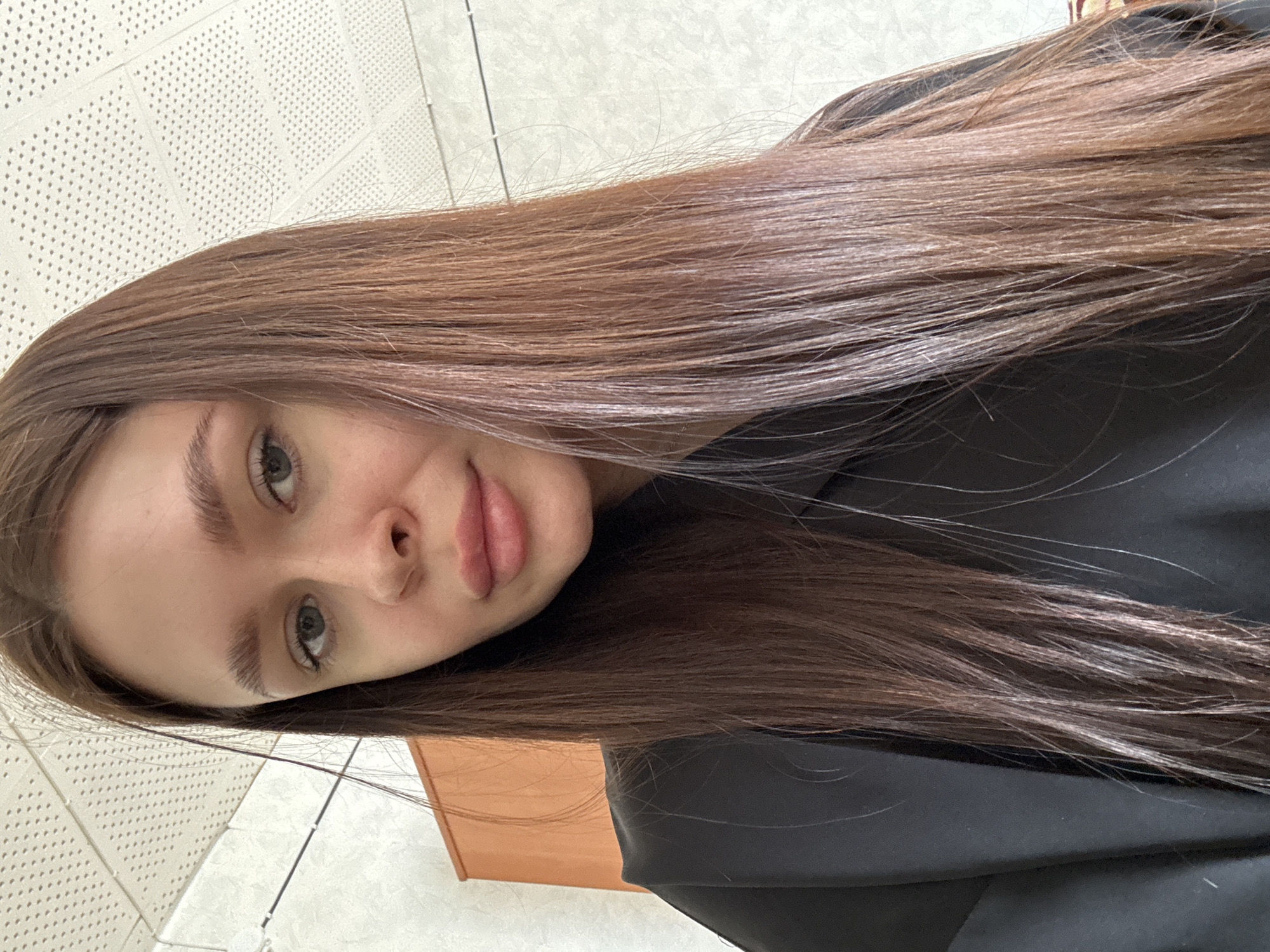 A young woman with long brown hair is centered in the frame. She is looking directly at the camera with a slight smile. She has light skin, grey eyes, and full lips. She is wearing a black garment. The background includes a wall with a textured, perforated surface and a light-colored tiled floor. Part of a wooden object is visible on the bottom left. There is no evidence in this image to indicate its location in Dubrova, Russia.