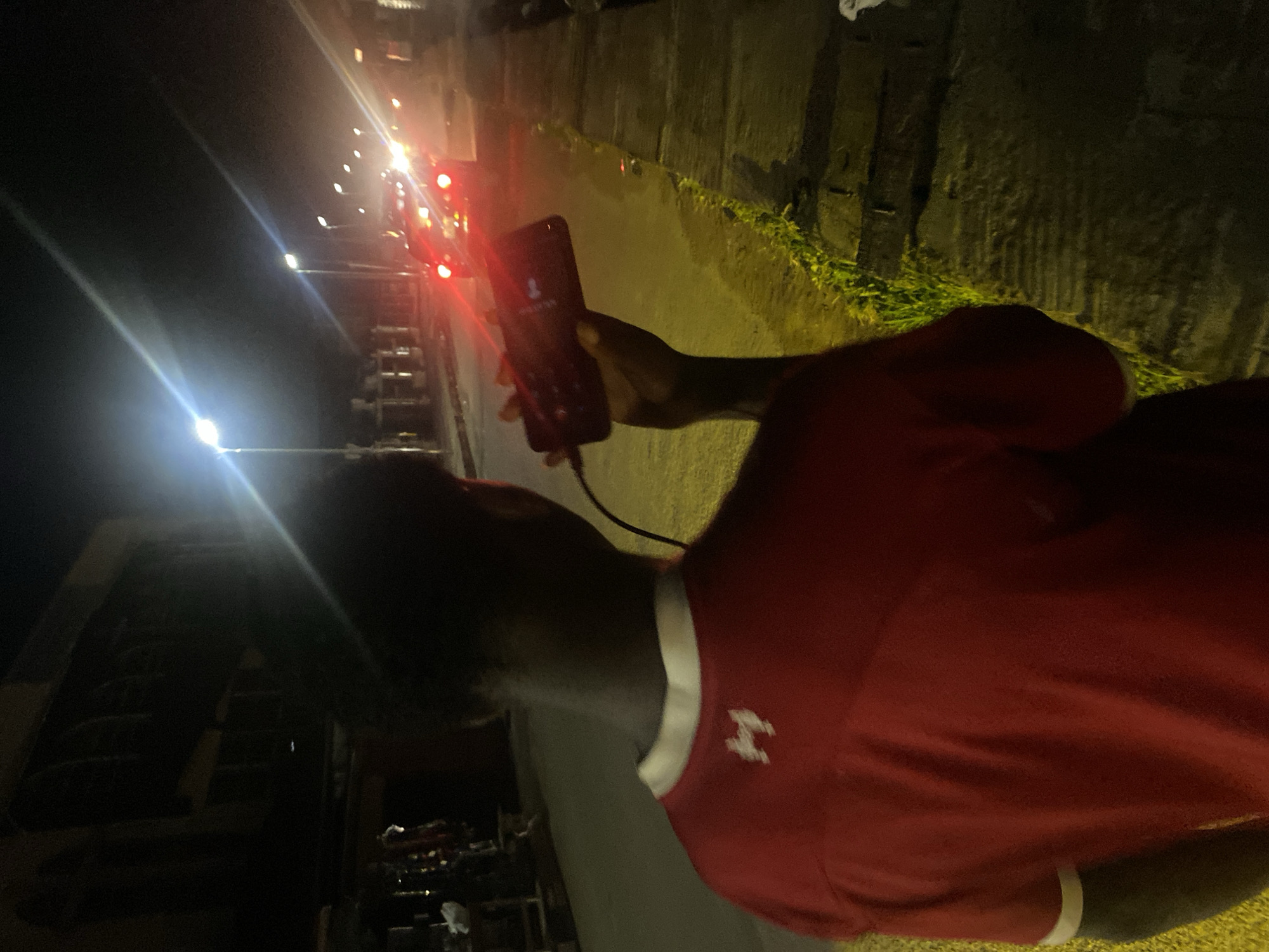 A person is holding a phone to their ear, seemingly on a call. The phone is black, and the screen is illuminated with what appears to be an incoming call interface. The person is wearing a red t-shirt with a white collar and white logo on the right side of the chest. The background shows a street scene at night with traffic lights and other vehicles. The scene has a slight overexposure, as if it was shot at night. Based on the background vegetation and the general appearance of the street, it is a general assumption that it is most likely the city of Lagos, Nigeria, but it is difficult to confirm the exact location without further information.