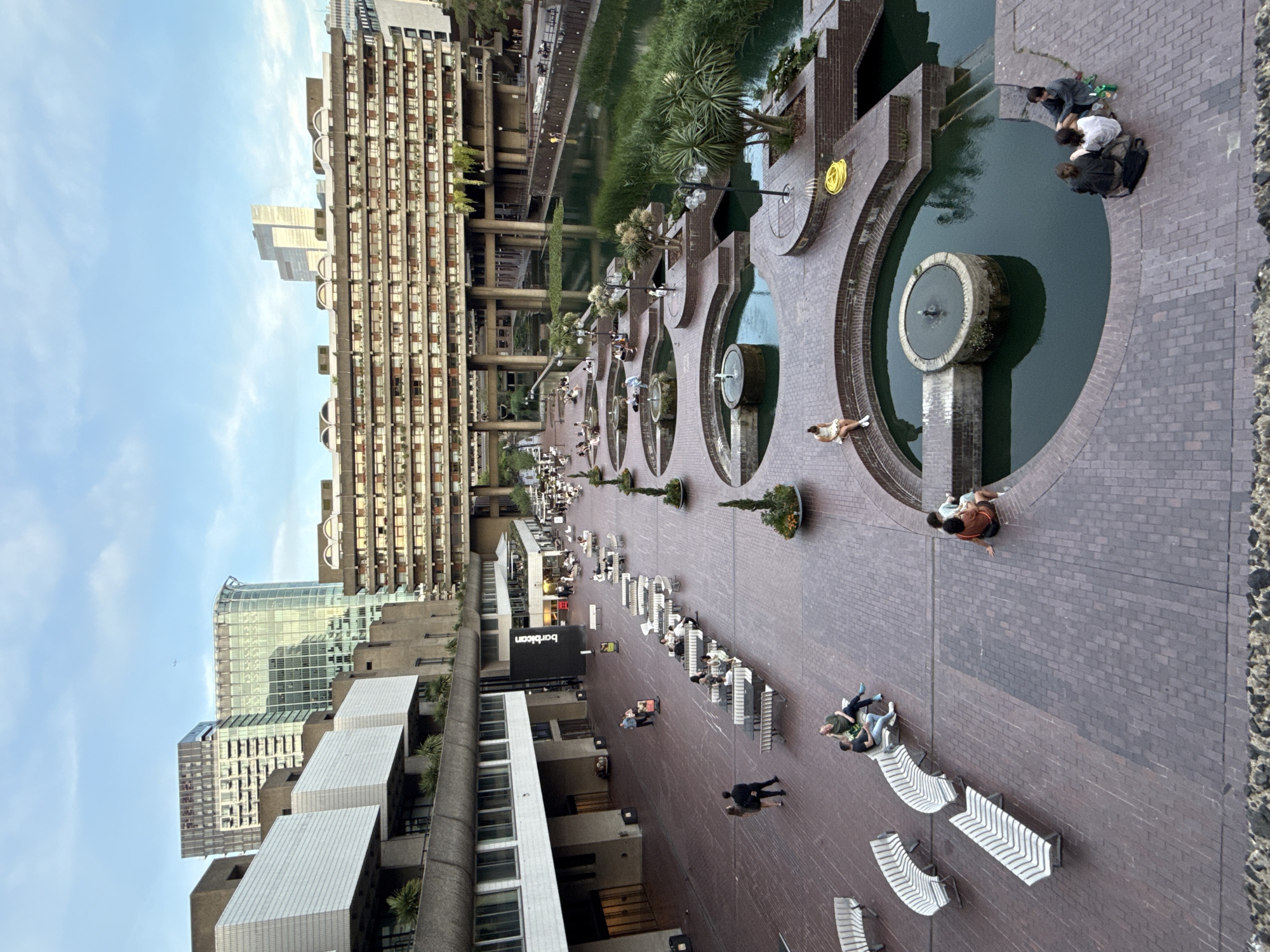 This high-angle shot depicts the Barbican Centre in London, UK. The architecture is characterized by brutalist buildings, concrete structures, and angular designs. A long pedestrian walkway, constructed of red bricks, stretches through the frame, lined with modern white benches where people are sitting and relaxing. Several people are walking along the path. Further down the walkway are a series of circular water features with fountains, each surrounded by greenery. The Barbican estate buildings rise along the left side of the walkway, while a large body of water with dense vegetation is situated on the right. The sky is partly cloudy, suggesting daytime, and various other buildings can be seen in the background.