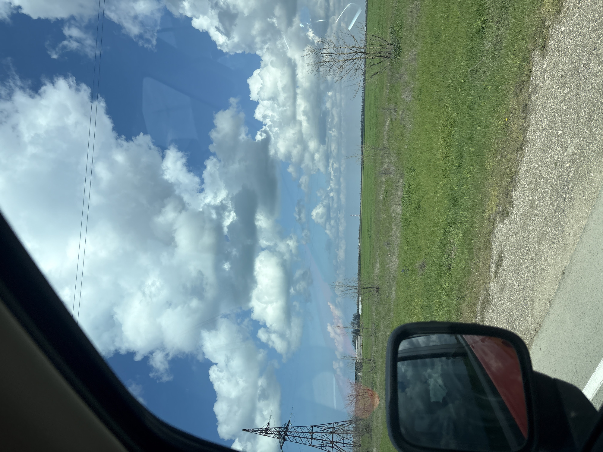 The image depicts a view from inside a vehicle, likely a car, in the Nikolaevskoe rural settlement area in Ukraine. The scene is oriented vertically.

Outside the window, there's a bright blue sky with numerous white, puffy clouds. A power transmission tower is visible near the bottom of the frame. A flat, green field stretches horizontally across the midground. Bare trees stand intermittently in the field. A road with a gravel shoulder runs along the right side of the frame.

The side mirror of the vehicle is visible in the lower right corner, reflecting the red color of the car's exterior. Part of the car's interior frame is also visible on the left side of the frame.
