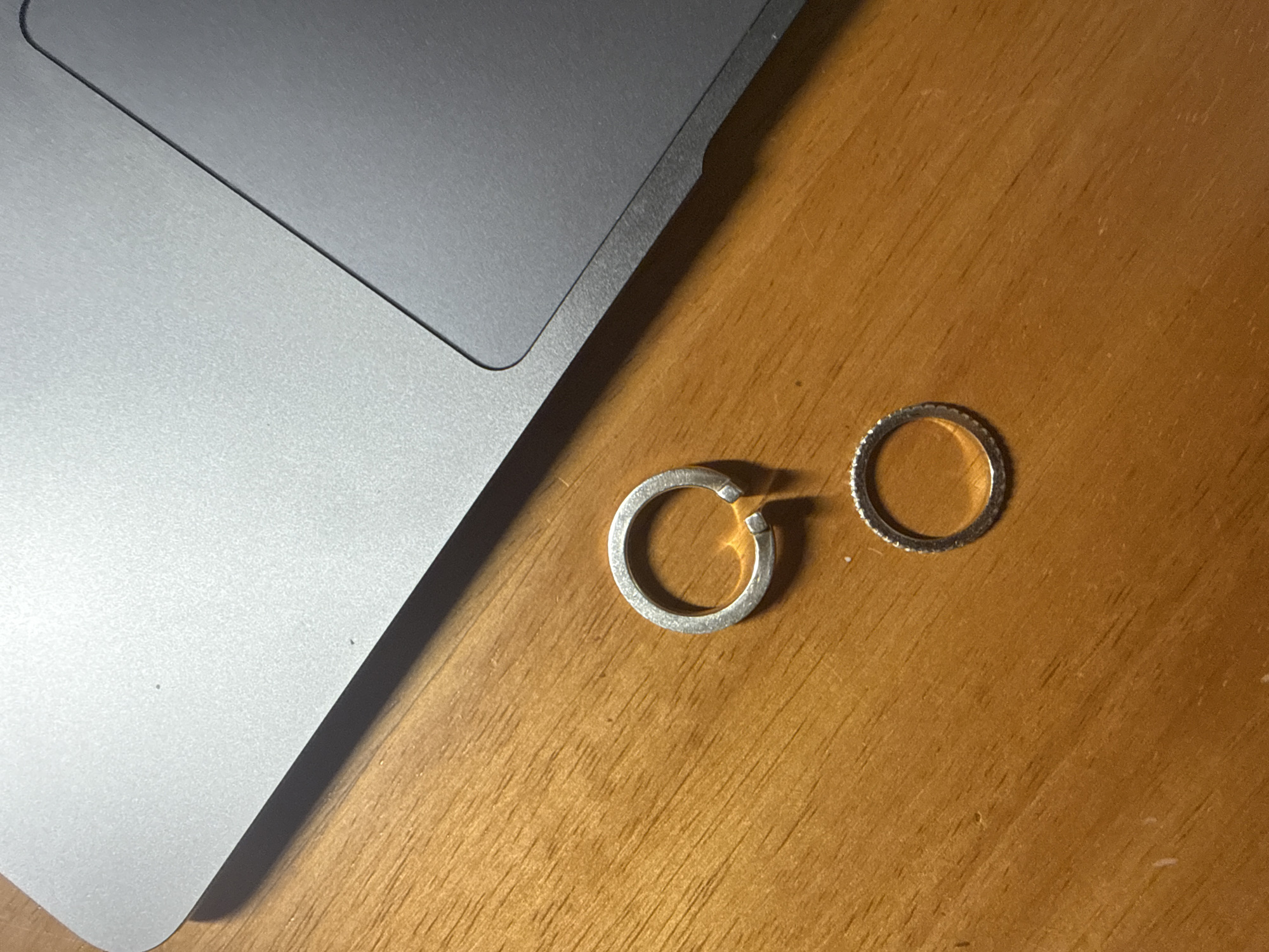 The image displays a close-up, overhead view of a silver-colored laptop and two silver rings resting on a wooden surface in San Bernardino Tlaxcalancingo, Mexico.
The top and left portions of the frame are occupied by part of an open laptop, revealing its metallic, matte-finished trackpad area and a section of the keyboard's base. A subtle seam or groove delineates different parts of the laptop's body.
To the right of the laptop, on a warm-toned wooden table, are two silver-colored rings. The ring on the left is open-ended, forming a C-shape, with a slightly textured or unpolished finish. The ring on the right is a complete circular band, featuring a distinct, finely ridged or studded pattern along its outer edge.
The wooden surface exhibits a visible grain pattern and some minor scuffs or imperfections. The lighting appears to be localized and warm, creating soft shadows and highlighting the textures of the objects, characteristic of an indoor setting, possibly under artificial light or in the evening. No individuals are present, and no specific activity is depicted. There is no visible text on any of the objects.