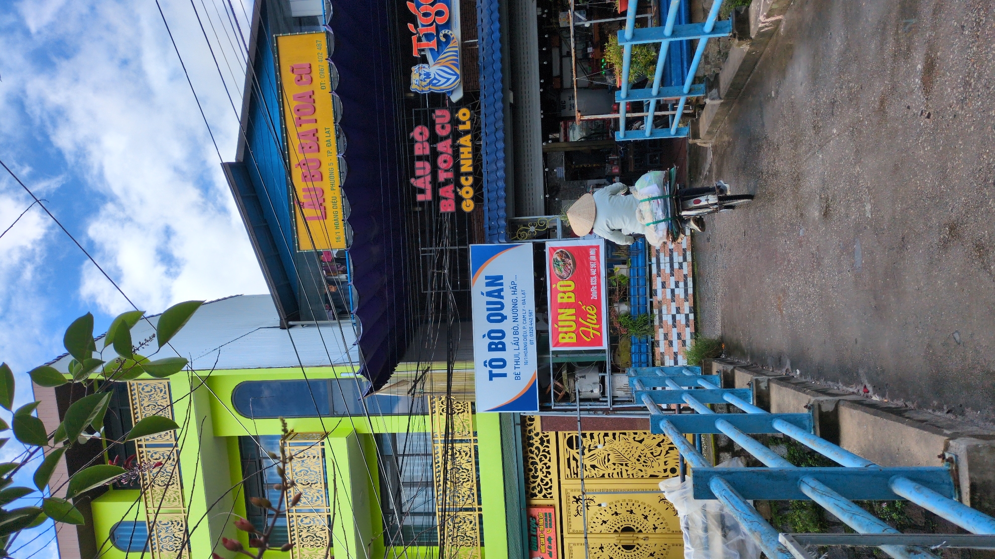 The image captures a street scene in Da Lat, Vietnam, focusing on a bridge with a bright blue railing. A person wearing a conical hat rides a bicycle loaded with goods across the bridge, heading away from the camera. The bicycle is covered with packages wrapped in plastic and secured with straps.
On the left side of the bridge, there is a multi-story building painted a vibrant lime green, featuring arched windows and decorative yellow railings on the balconies. Adjacent to it is a blue awning and a yellow sign with Vietnamese text that reads "LẨU BÒ BA TOA CŨ GÓC NHÀ LÒ" and "Tiger", indicating a restaurant or food establishment. Below this, a blue and white sign for "TÔ BÒ QUÁN" is visible, advertising "Bò thui, Lẩu bò, nướng, hấp..." with an address and phone number. Another smaller sign below that prominently displays "BÚN BÒ Huế" with a phone number.
On the right side of the bridge, there are more storefronts with awnings. The ground surface of the bridge and the surrounding street appears wet, suggesting recent rain. The overall environment depicts a bustling urban setting with signs of commercial activity, typical of a Vietnamese city. The sky is partly cloudy, with patches of blue.