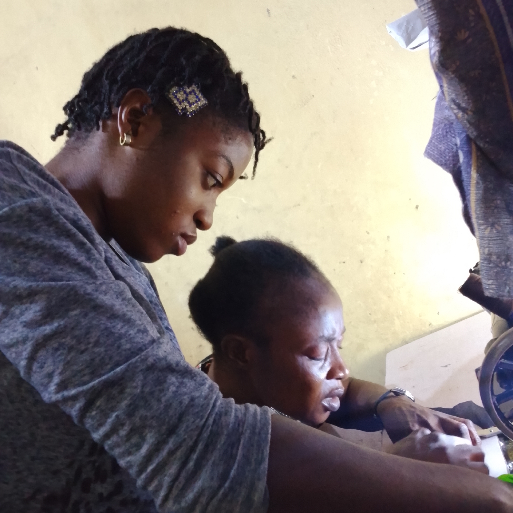 This is a medium shot, indoor, eye-level photograph depicting two Black women in a dimly lit room, possibly in Ado, Nigeria. The woman in the foreground is on the left, wearing a grey, textured long-sleeved shirt. Her head is turned to her right, and she is looking down at the woman in the background. She has dark, braided hair adorned with a decorative clip featuring blue and gold geometric shapes. A small gold hoop earring is visible in her left ear.
The second woman is in the background, her face partially obscured and turned to her right, with her eyes closed. She is wearing a dark top, and her hair is pulled back into a bun. Her expression appears pained or distressed, with her mouth open. Her right hand is visible, and she is touching her face.
The environment is sparse, with a yellow-painted wall in the background. A patterned textile, possibly a curtain or blanket, hangs on the right side of the frame. A portion of a sewing machine's wheel and part of a white surface, possibly a table, are also visible on the right. The lighting is soft and appears to be coming from an artificial source. The overall mood of the image is somber and intimate.