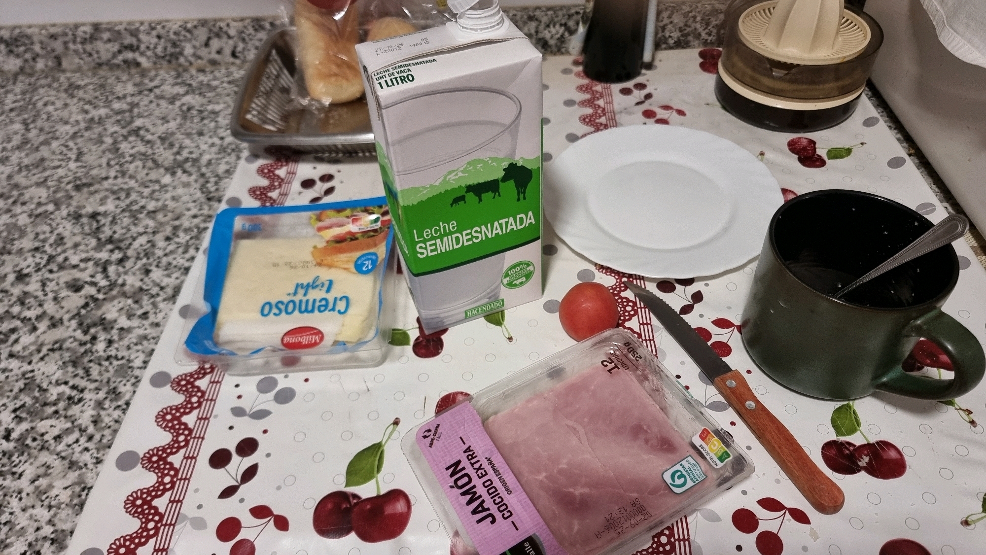 The image depicts a table set for breakfast in Sant Joan Les Fonts, Spain. The scene includes a carton of semi-skimmed milk, a package of sliced cheese, a package of cooked ham, a small tomato, a white plate, a dark green mug with a spoon inside, and a knife with a wooden handle. In the background, there is a tray with bread and a manual juicer. The table is covered with a tablecloth featuring a cherry pattern. The lighting suggests it is daytime.