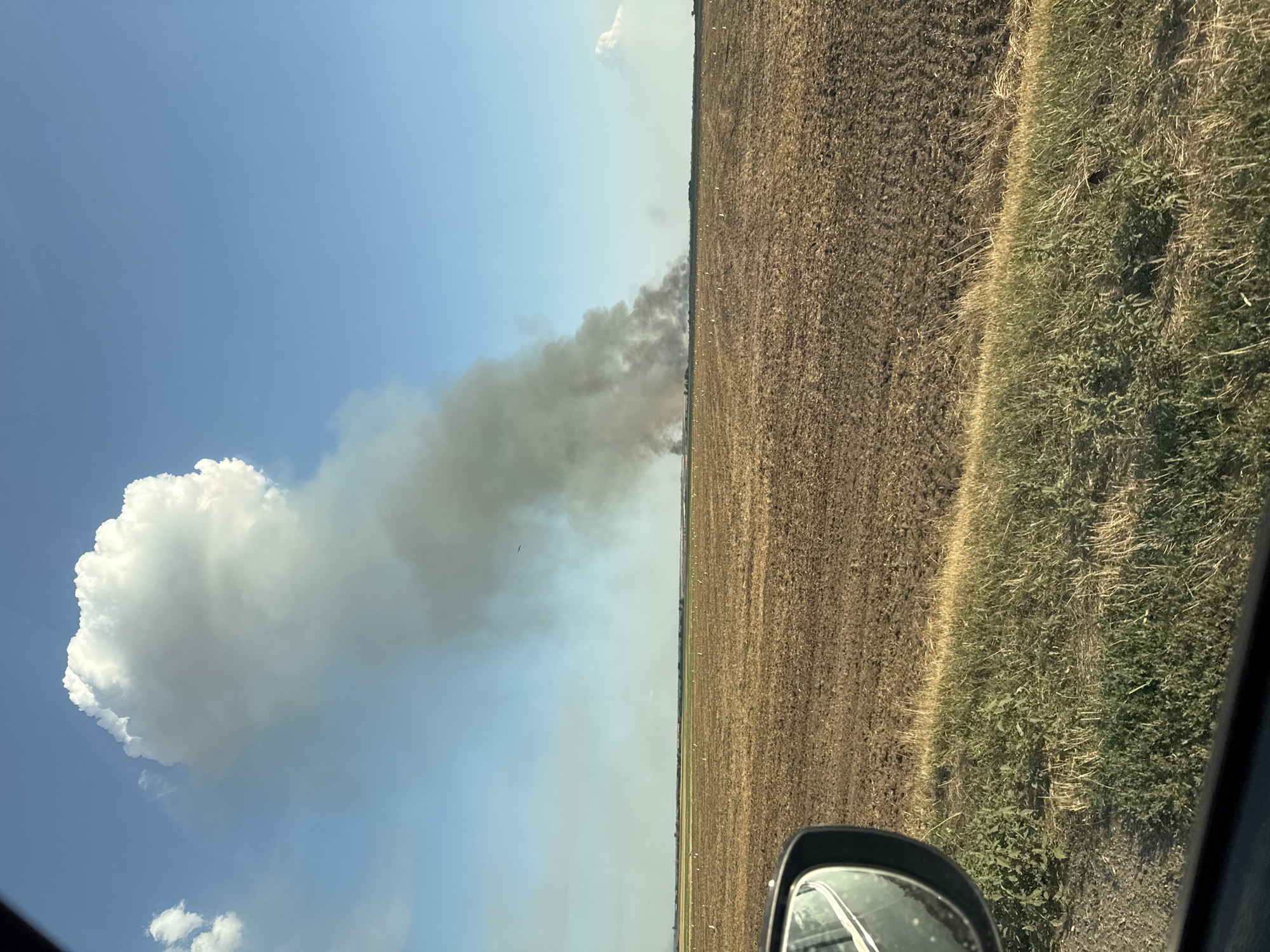 The image depicts a rural landscape under a clear blue sky with a large, billowing cloud of dark smoke rising from the distance. The smoke plume is extensive and appears to originate from a fire on the horizon. In the foreground, there is a field of dry, tilled earth, with patches of green grass and vegetation along the bottom edge of the frame. A car's side mirror is visible in the lower left corner, suggesting the photo was taken from inside a vehicle. The overall scene conveys a sense of an ongoing environmental event, likely a large fire. The location of Zhukovskoe, Russia, is not discernible from the visual information provided in the image.
