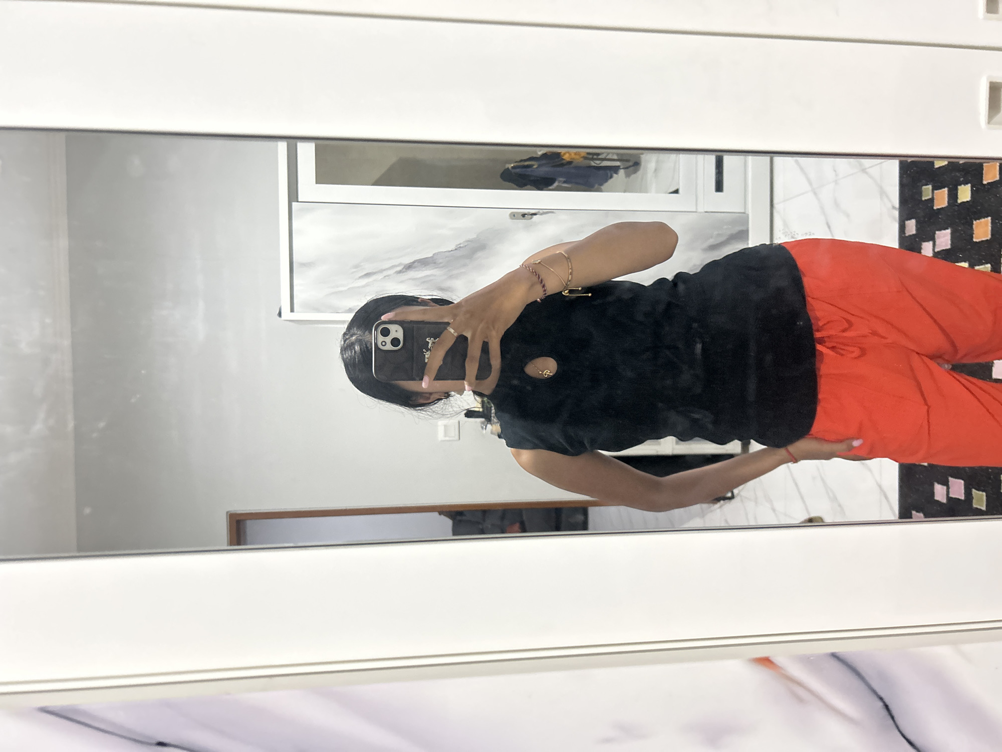 A woman is taking a mirror selfie in a room. She is wearing a black sleeveless top and bright orange pants. She is holding a smartphone in her right hand to take the photo. The room appears to be a bedroom with a large white wardrobe with reflective doors and a marble-patterned wall in the background. A portion of a framed artwork with an abstract design is visible on the left side of the image. The flooring is tiled with a marble pattern. The location is identified as Sukawati, Indonesia.