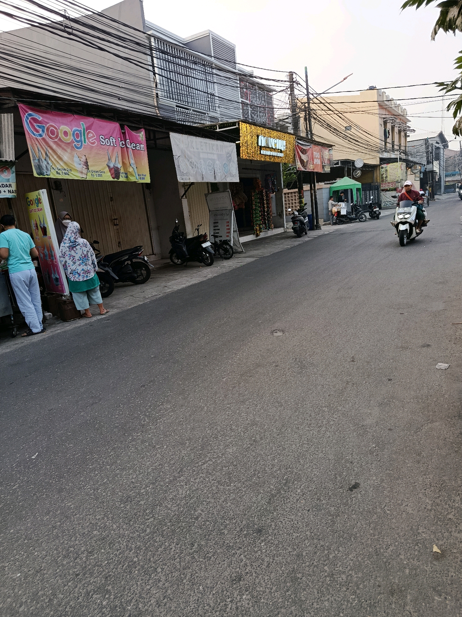 The image depicts a street scene in Bekasi, Indonesia, with a focus on a row of shops and passing traffic. In the foreground, a paved road occupies the majority of the frame, showing signs of wear. On the left side of the road, a sidewalk area features several individuals and parked motorcycles. Two people, one in a blue shirt and white pants, and another in a patterned hijab and green pants, appear to be customers or employees near a stall displaying ice cream cones with a "Google Soft Ice Cream" banner. Several motorcycles are parked along the sidewalk.
Further down the street, more shops are visible, identifiable by signage. One shop has a bright, gold-colored facade with the word "noveus" illuminated. Another sign reads "TOKO HYA." A green tent structure is set up on the sidewalk, with a few individuals and motorcycles nearby.
In the middle ground, traffic is present. A white scooter carrying two individuals is moving away from the camera, heading down the street. The street itself is lined with buildings, and a complex network of electrical wires is suspended above the scene, a common sight in urban areas of Indonesia. The architecture of the buildings appears to be a mix of modern and older structures. The overall lighting suggests it is either early morning or late afternoon, with diffused sunlight. The environment appears to be a busy commercial area within Bekasi.