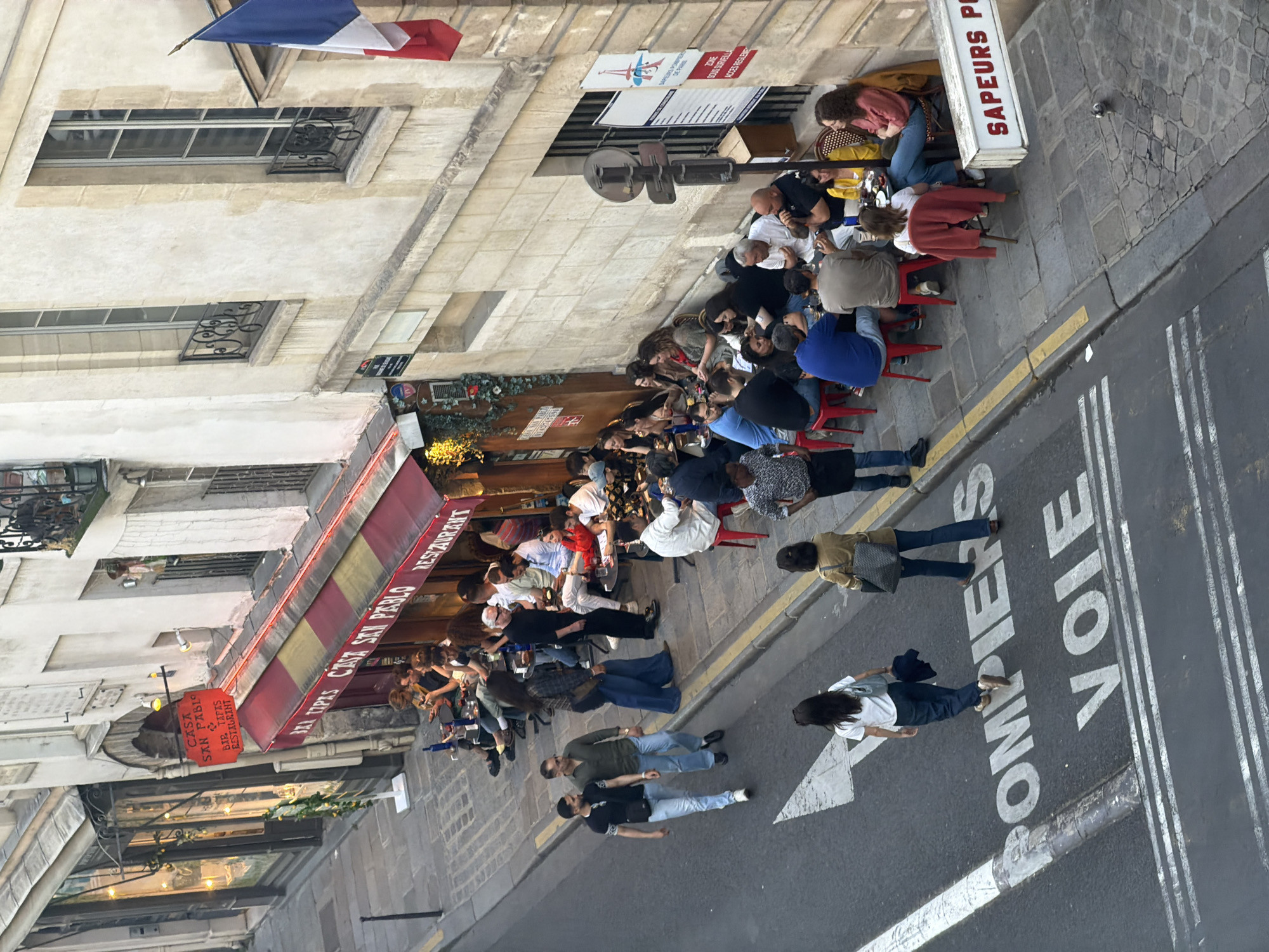 The image shows an outdoor scene in Paris, France. A restaurant called "Casa San Pablo" is visible with a red and yellow awning and a sign indicating it's a bar, tapas restaurant. People are seated at tables outside the restaurant, eating and socializing. Pedestrians are walking on the street next to the restaurant, where a painted arrow and the word "POMPIERS" (Firefighters) and "VOIE" (Way) are visible, indicating a fire lane. A French flag is displayed above a building in the background. A sign marked SAPEURS POMPIERS (firefighters) can be seen adjacent to the diners.