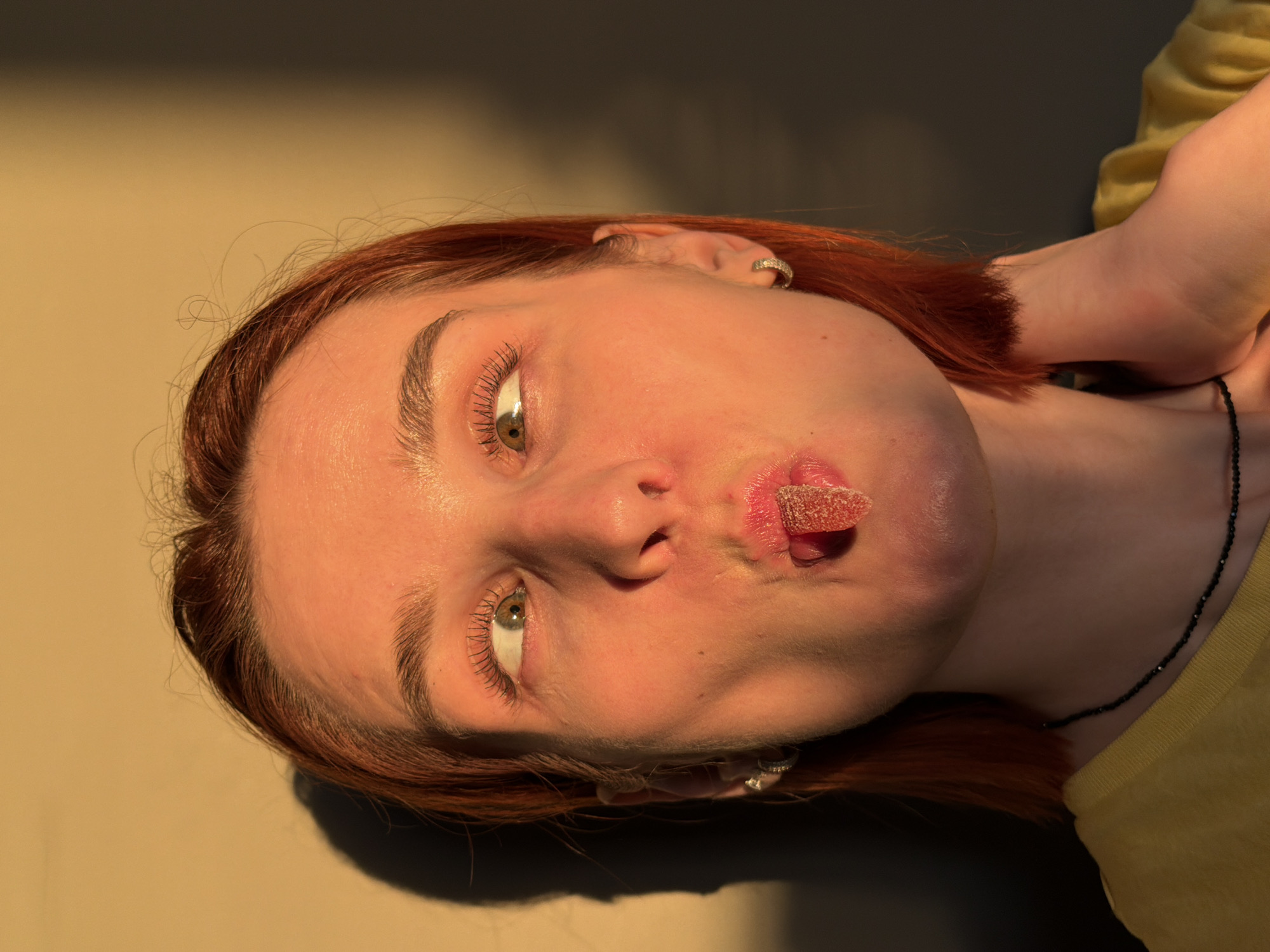 The image shows a woman lying down, with her head resting on a surface. She has red hair and light skin. She is wearing a yellow top and a black necklace. She has a red gummy candy in her mouth. The lighting is warm and casts shadows across her face and the background. The background appears to be a plain, neutral color. The location is Chaikovsky, Russia.