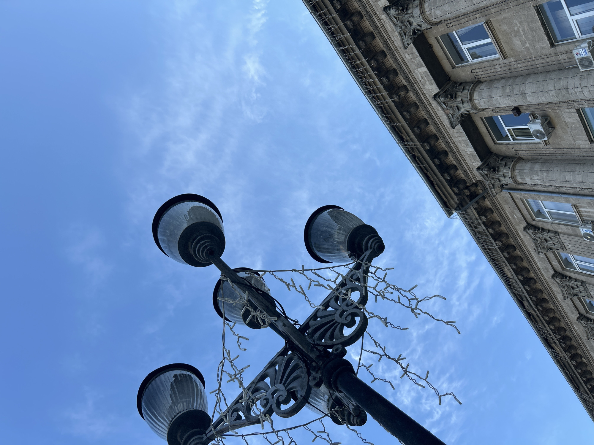 The image shows a street lamp with multiple globes, adorned with string lights, against a blue sky with thin clouds. To the right, a section of an old building with classical architectural elements, including columns and ornate detailing above the windows, is visible. The building appears to be made of stone and has several windows, some with air conditioning units installed. The photograph is taken from a low angle, looking upwards. The specific location is the city of Bălți, Moldova.