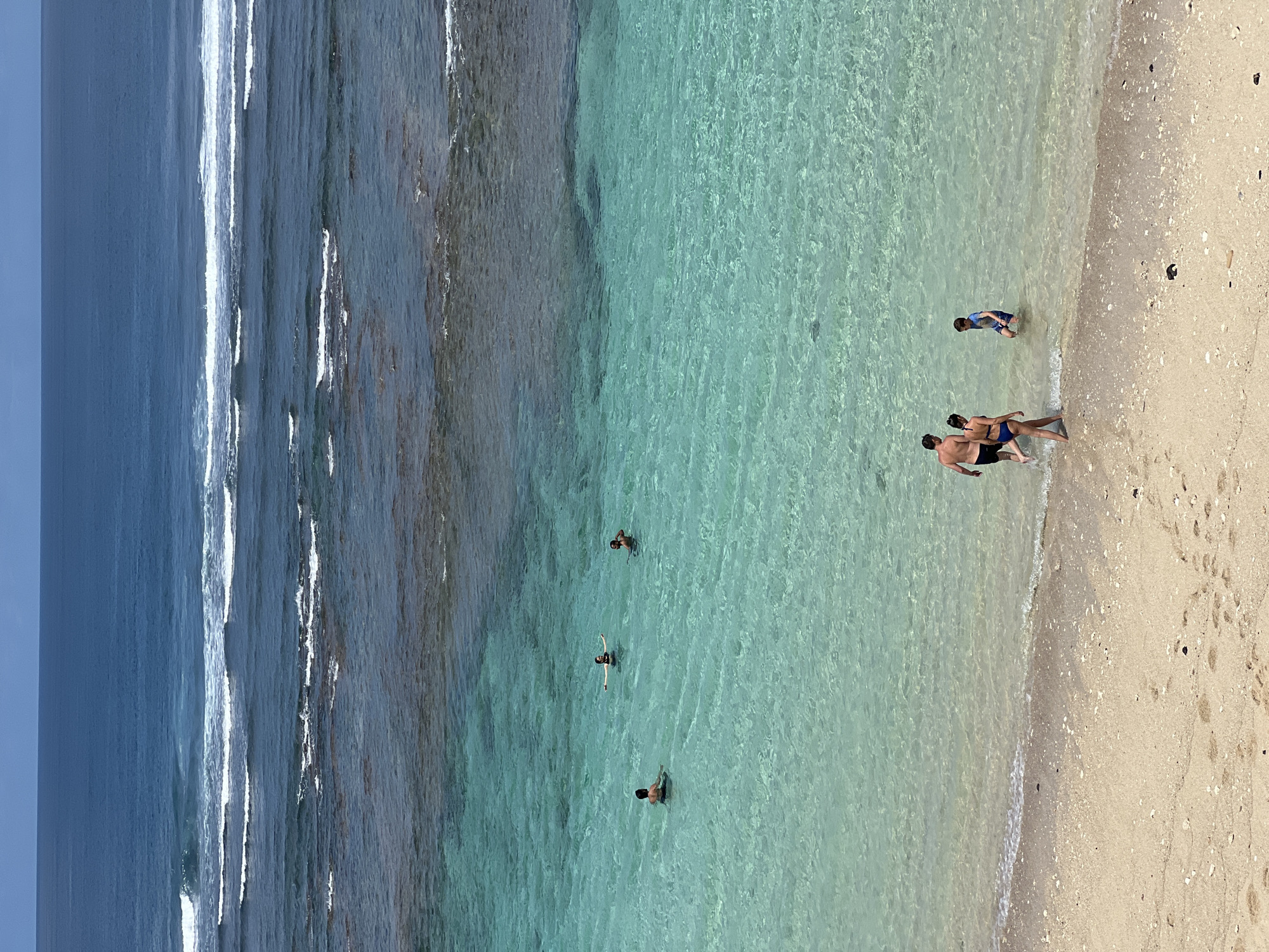 This image presents an elevated view of a vibrant beach scene in Kuta Selatan, Indonesia, showcasing clear turquoise waters meeting a light sandy shore under bright daylight.
The main subjects are several people enjoying the ocean. In the foreground, near the shoreline, a man and a woman, both in swimwear, stand partially submerged in the transparent shallow water. A bit further out, a child, clad in a blue shirt and dark shorts, wades in the remarkably clear water. Deeper into the water, at least three more individuals are visible; one person is floating on their back, another is standing or wading, and a third figure is less distinct.
The scene transitions from the pale, shell-strewn sand on the right, which shows faint footprints, to the crystal-clear, shallow turquoise water where the people are. Further left, the water deepens into a darker blue, characterized by white-crested waves rolling in from the open sea. The waves near the shore are gentle.
The time of day appears to be bright and sunny, indicative of ideal beach weather. The atmosphere is calm and recreational. There are no visible signs of text in the image.