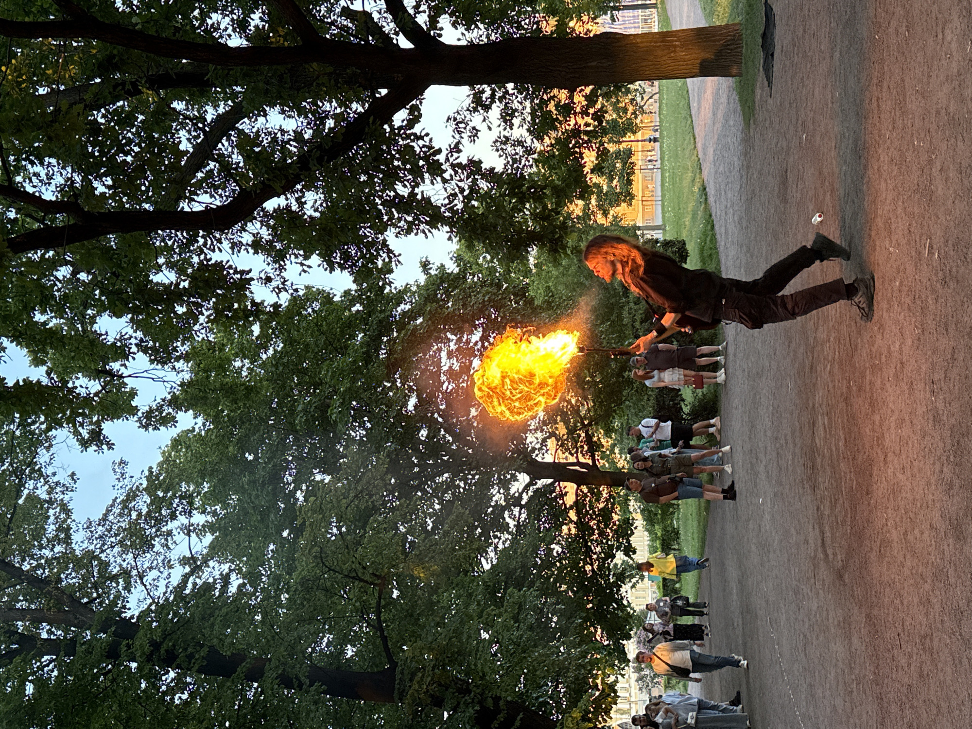 An outdoor scene at dusk in a park in Saint Petersburg, Russia, features an individual performing fire-breathing. The performer, appearing male with long hair and dark clothing (long-sleeved shirt, cargo pants, athletic shoes), is positioned towards the right of the frame, leaning back with knees bent, expelling a large, bright orange and yellow ball of flame from his mouth. He holds a dark, slender torch-like object in his right hand, angled upwards.
Numerous mature trees with dense green foliage dominate the upper and lateral portions of the image, allowing patches of light blue twilight sky to be visible. A dirt or gravel pathway occupies the foreground and midground, where multiple onlookers are present. On the left, a group of individuals walks towards the viewer. In the mid-ground, a cluster of approximately ten people stands, observing the fire performance; one person among them wears a distinct bright yellow jacket. Distant light-colored buildings with classical architectural details are discernible through the trees in the background, particularly on the left side, suggesting an urban environment. The ground underfoot transitions from the pathway to grassy areas further back. A small, white cylindrical object rests on the path near the performer's left foot, and a dark cloth lies on the grass by a tree trunk on the right. The overall lighting suggests the fading light of evening, with the fire providing the most intense illumination.