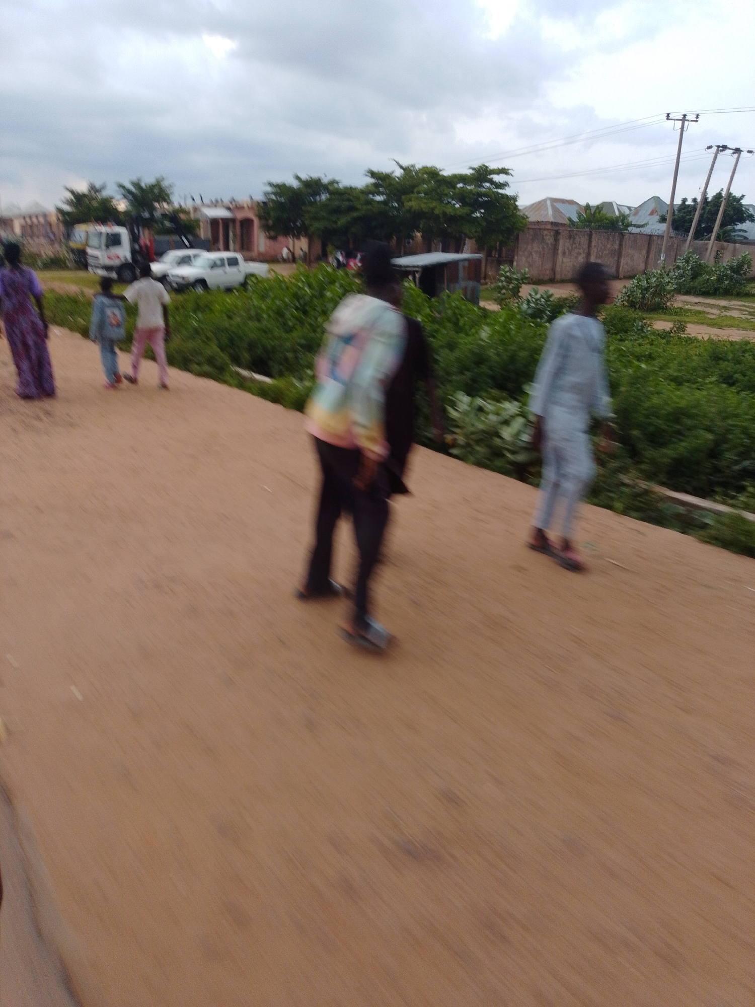 The image depicts a scene on a dirt road in Gombe, Nigeria, under an overcast sky. Several individuals are walking along the road. In the background, there are buildings with corrugated metal roofs and some vegetation. A white truck and a white car are visible near the buildings. Power lines and poles are also present. The image appears to have been taken while the camera was in motion, resulting in motion blur throughout the scene.