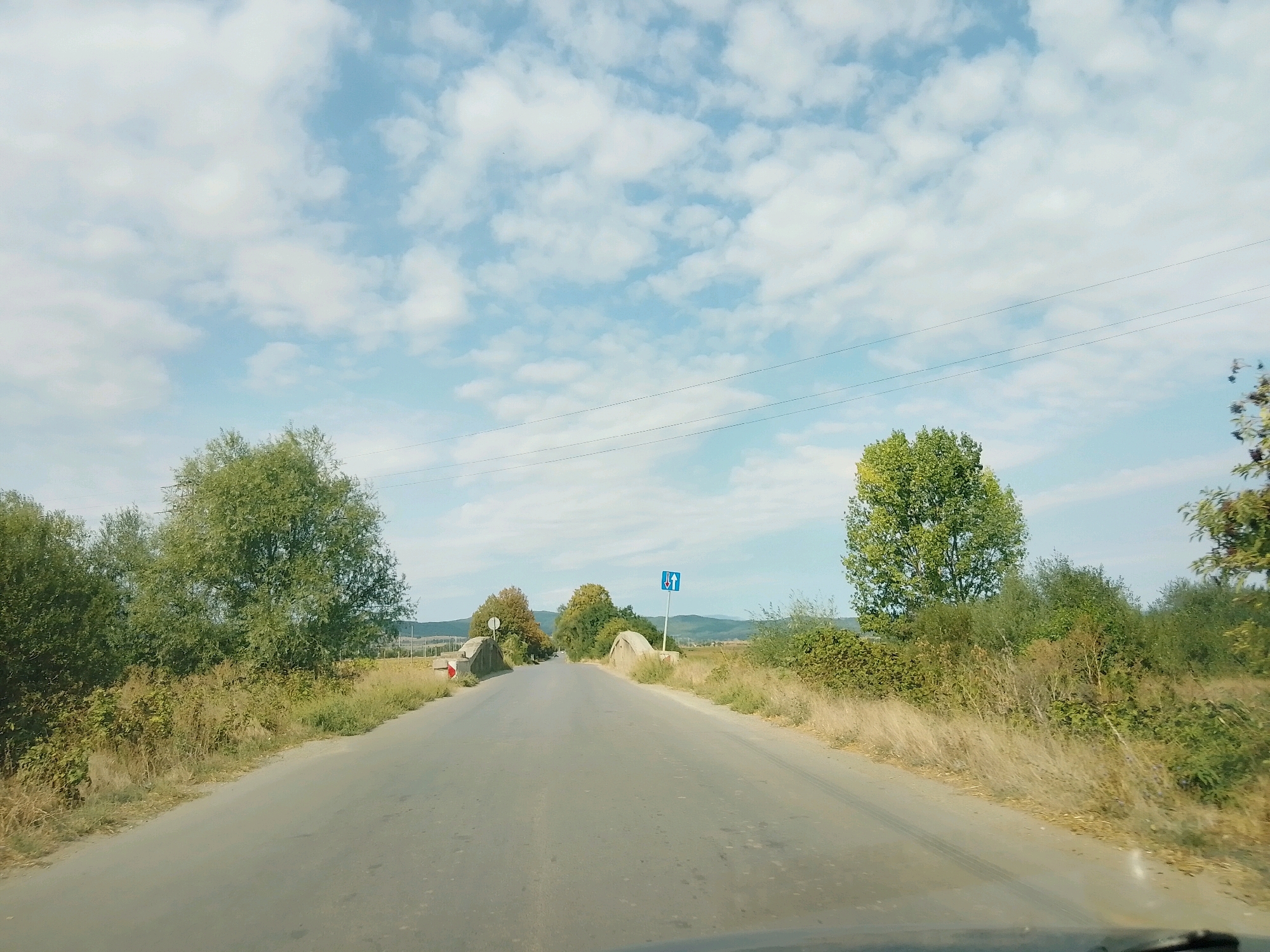 The image shows a two-lane asphalt road passing through a rural landscape. On either side of the road are overgrown grass and bushes, with several trees scattered throughout. In the distance, rolling hills are visible under a partly cloudy sky. The road appears to lead towards a small bridge or overpass. A blue traffic sign with white arrows indicating two-way traffic is posted on the right side of the road. There are no people or vehicles visible in the image. The scene suggests a remote, possibly unpopulated area.