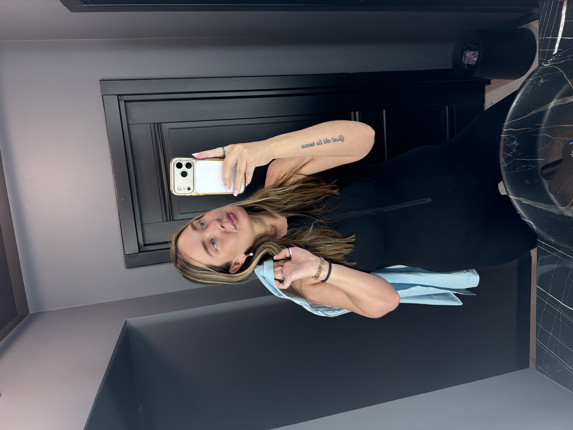 A woman is taking a selfie in a bathroom. She is holding a cell phone in her right hand. She is wearing a black dress and has a light blue towel draped over her left arm. The bathroom has gray walls and a black door. There is a black waste bin and a marble sink in the background. The location is not discernible as being in Moscow, Russia.