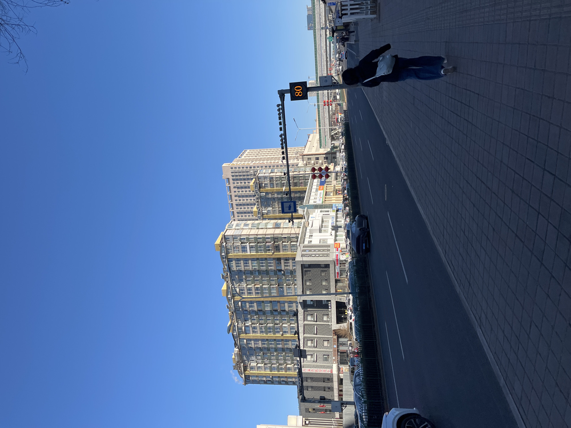 The image depicts a street scene in Changchun City, China. A person is walking on the sidewalk adjacent to a multi-lane road. Several vehicles are present on the road. A tall building with multiple windows and yellow detailing is visible, situated behind a traffic signal displaying the number "80". The sky is clear and blue.