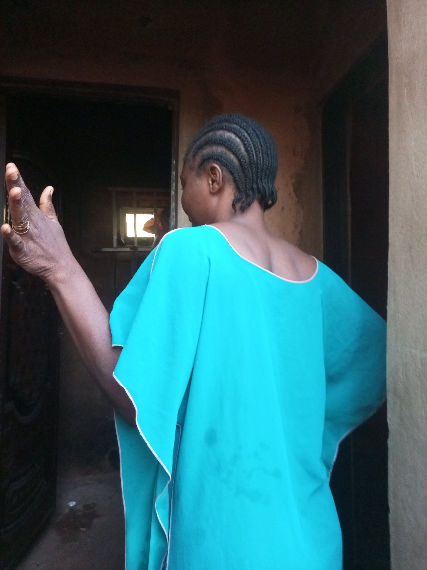 The image shows a person from behind, facing a doorway. The person has dark hair braided in cornrows and is wearing a bright turquoise garment with a light pink trim around the neckline. Their right arm is raised, with the hand open and facing forward. A gold-colored ring is visible on the index finger of this hand. The doorway is dark and appears to lead into a building. Through the doorway, a window with a white frame is visible, and light is coming from outside. The walls around the doorway are a textured, earthy brown color. The scene appears to be outdoors or in an open doorway in a rustic setting. The location is not specified as Tempure, Nigeria.