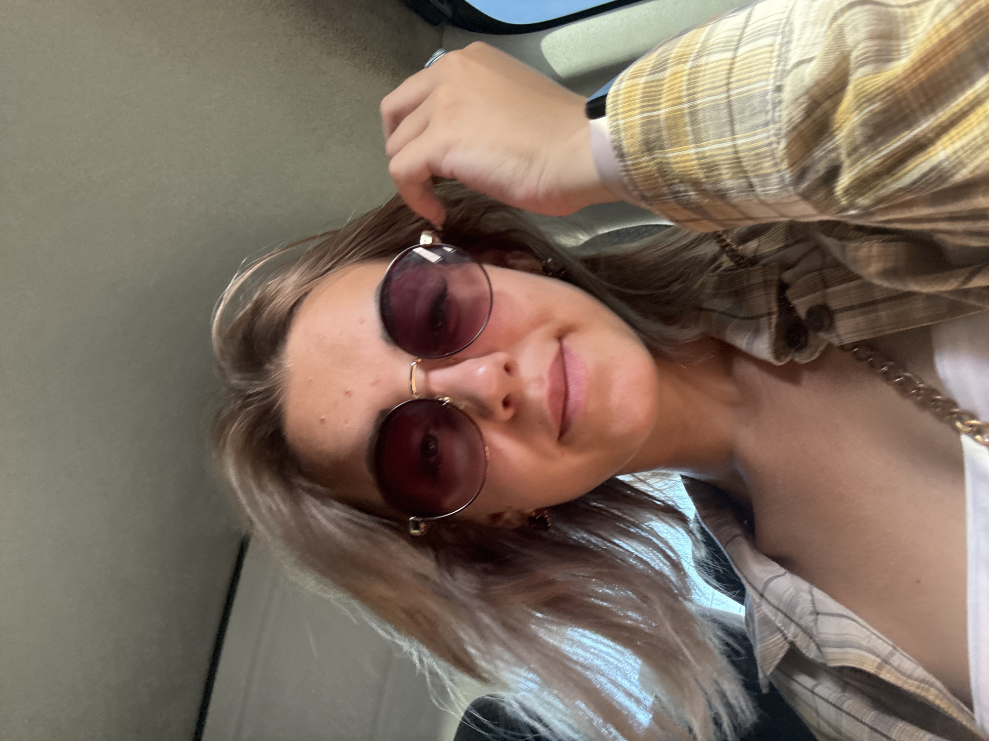 A young woman with blonde hair and sunglasses is in the back seat of a vehicle. She is wearing a plaid shirt and a white top. Her hand is on her sunglasses. The environment appears to be daytime and possibly sunny.