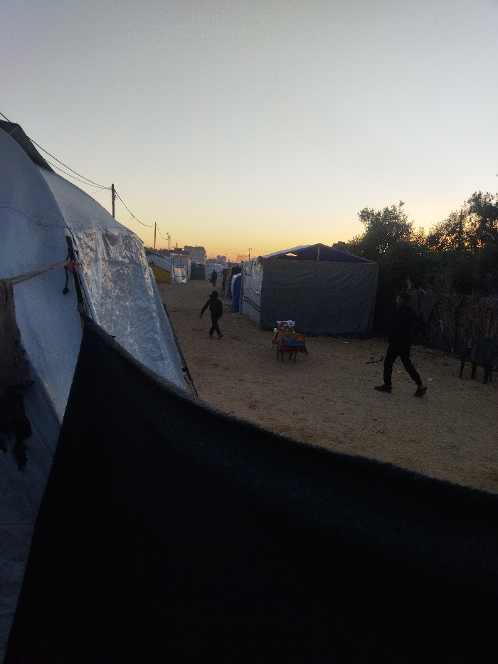 The image depicts an informal settlement in the city of Khan Yunis, Palestinian Territory, during either sunrise or sunset, with the sky displaying a gradient from light grey-blue overhead to orange-yellow at the horizon. The foreground features a large, grey tent-like structure, constructed from sheeting and secured with ropes, partially obscured by a dark fabric in the lower left. A wide, unpaved dirt pathway extends into the distance, flanked on both sides by numerous makeshift shelters crafted from various tarpaulins and fabrics.
In the midground, a person dressed in dark clothing walks away from the viewer towards the left side of the path. Another person, also in dark attire, walks towards the right side of the path, moving away from the viewer. Further along the pathway, several indistinct figures are visible. Near the right side of the path in the midground, a small, low, multi-colored table or stand is present, with two dark chairs positioned beside it. Utility poles supporting power lines are visible along the path in the mid-distance. Sparse vegetation, including trees or bushes, is situated on the right side of the scene, behind some of the shelters. Distant urban buildings are silhouetted against the glowing horizon in the background.