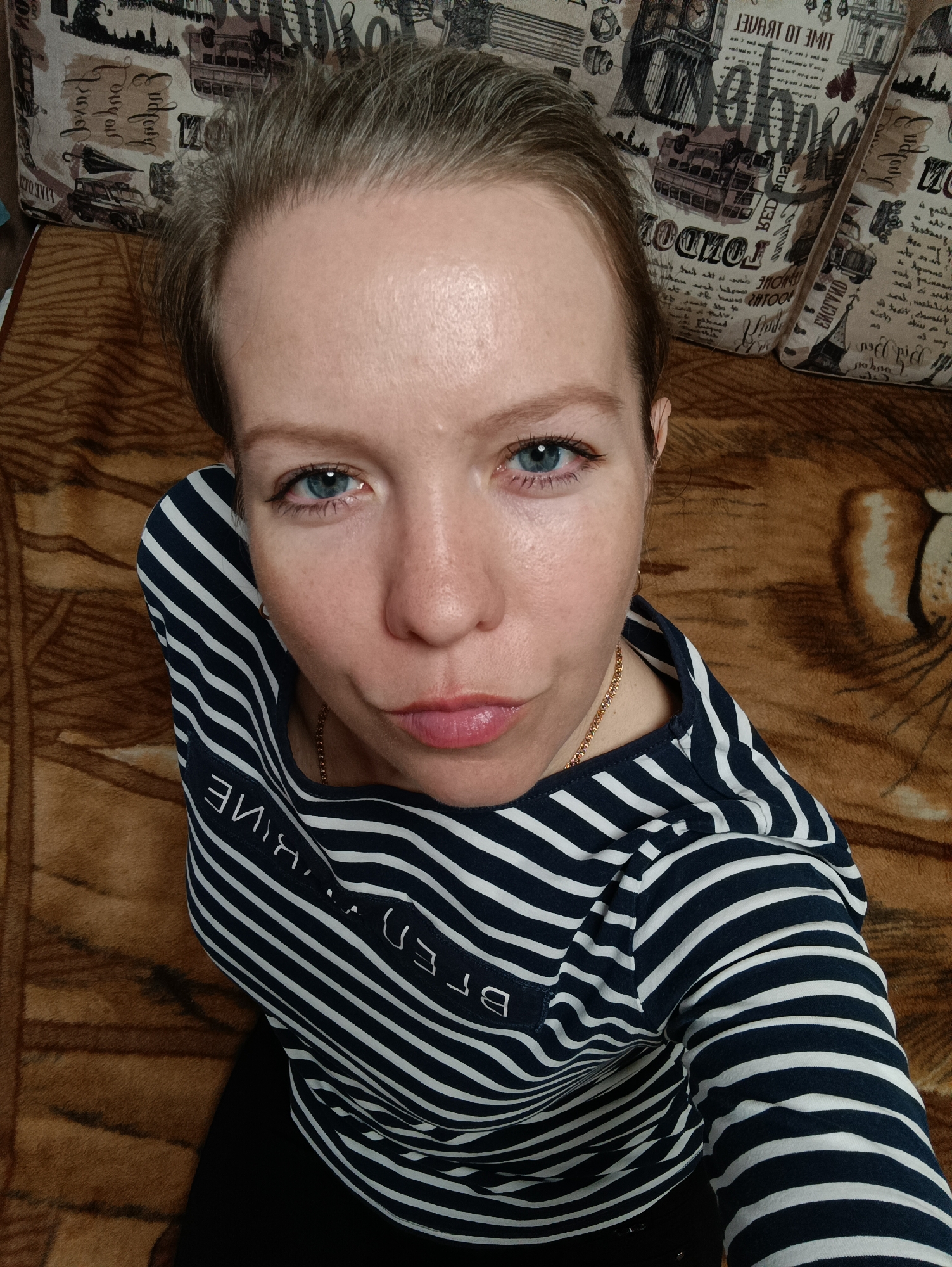 The image shows an overhead selfie of a woman with light skin, fair complexion, and striking blue eyes, taken indoors in Chaikovsky, Russia. She is looking directly at the camera with a subtle pouting expression. Her light brown hair is pulled back, and her forehead appears slightly shiny.
She is wearing a navy blue and white striped long-sleeve shirt, typical of a marinière style. The word "BRETON" is visible in white letters on the left sleeve, appearing reversed in the image. A delicate gold chain can be seen around her neck. She is seated or reclining on a warm brown, textured blanket or throw, which features a subtle animal print, with what appears to be part of a tiger's face visible on the right side of the frame.
Behind her, two pillows are visible, covered in a fabric patterned with various travel-related illustrations and text, predominantly featuring London landmarks such as Big Ben and a double-decker bus. Recognizable (though reversed) text on the pillows includes "LONDON", "TIME TO TRAVEL", "GUIDE", and fragments like "RED ROVER". The lighting suggests ambient indoor conditions. The overall impression is a casual, direct portrait with a hint of playfulness.