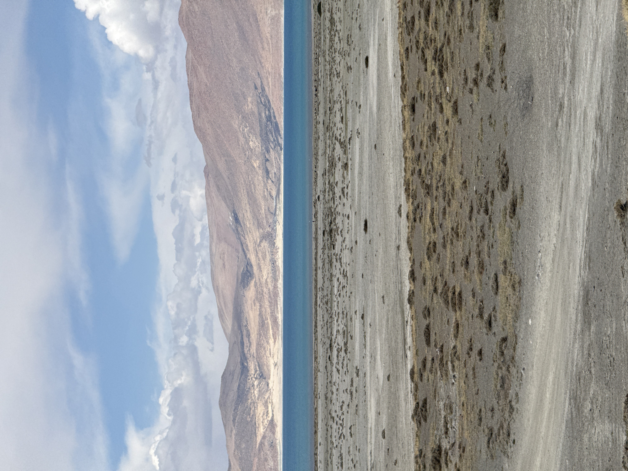 The image depicts a landscape with a mountainous region on the left, a body of water (possibly a lake) in the center, and a barren or arid land area on the right. The sky with clouds is visible above the mountains. The color palette is dominated by earthen tones, blues, and grays. The scene suggests a remote, high-altitude environment, potentially located in Gyirong County, China. There are no visible people, clear subjects, or objects.