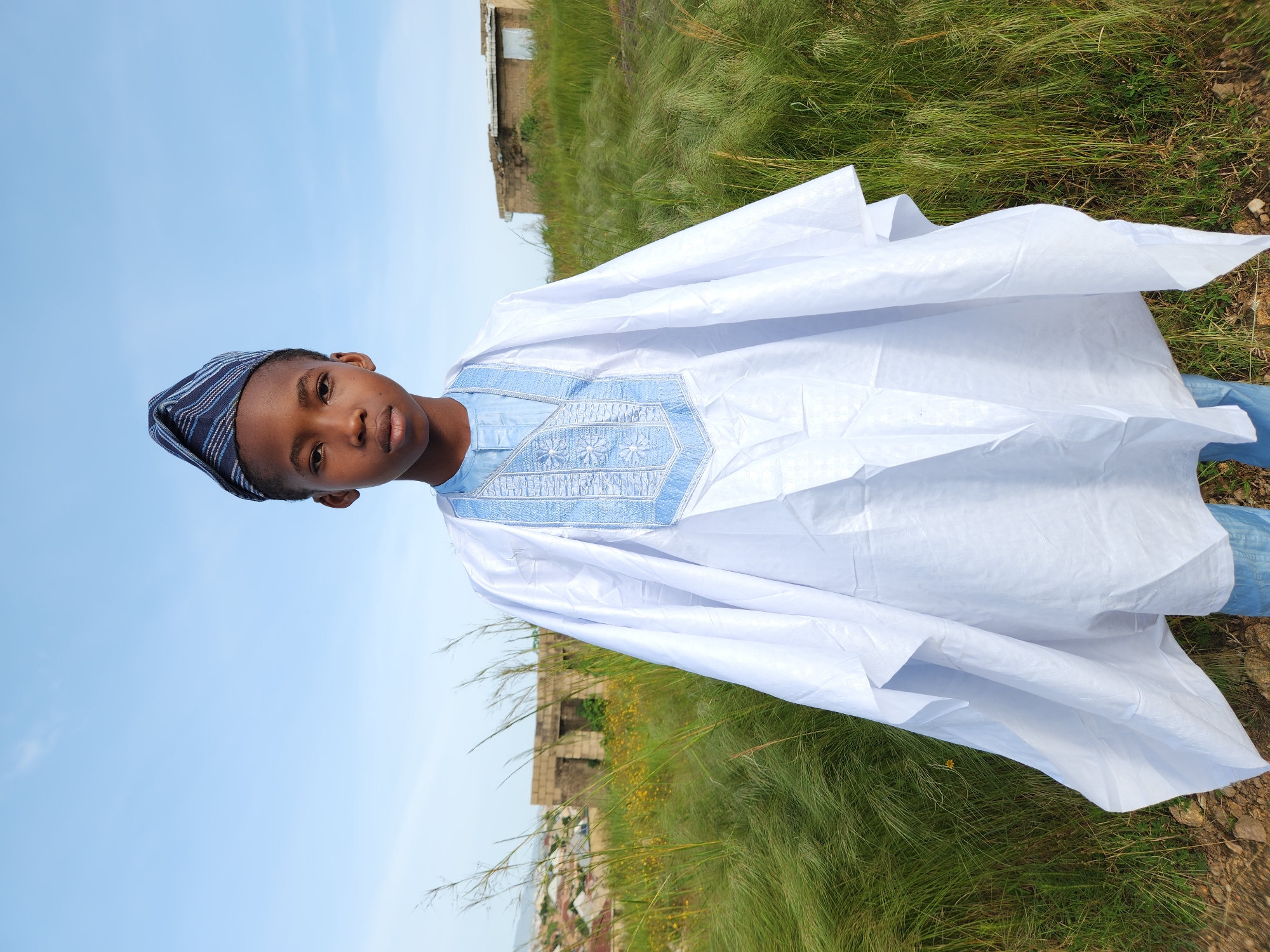 A young boy stands in the foreground, looking directly at the camera. He is wearing a white traditional outfit with light blue embroidery on the chest and a matching blue and white striped cap. The boy is positioned in an outdoor setting with tall, dry grass and some yellow wildflowers. In the background, there are dilapidated buildings and the distant outline of hills, suggestive of an undeveloped or rural area. The sky above is a clear blue with a few wisps of clouds. The scene appears to be set in the city of Jos, Nigeria.