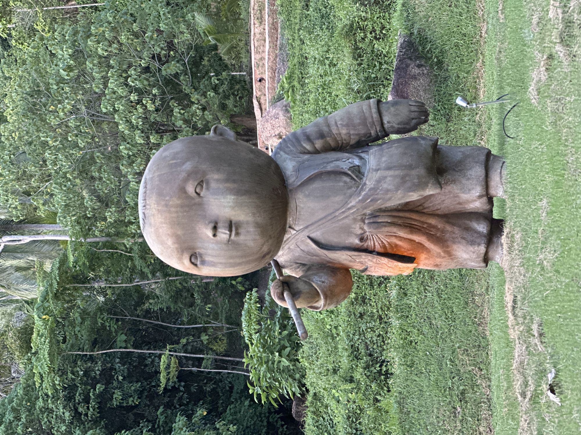 A bronze statue of a child with a large head, wearing a tunic and holding a stick, stands in a grassy area. The statue has a serene expression and appears to be illuminated from within by an orange glow. Behind the statue, lush green trees and foliage create a dense background. In the foreground, a small metal spotlight is planted in the grass. The scene is located in Baan Lipa Noi, Thailand.