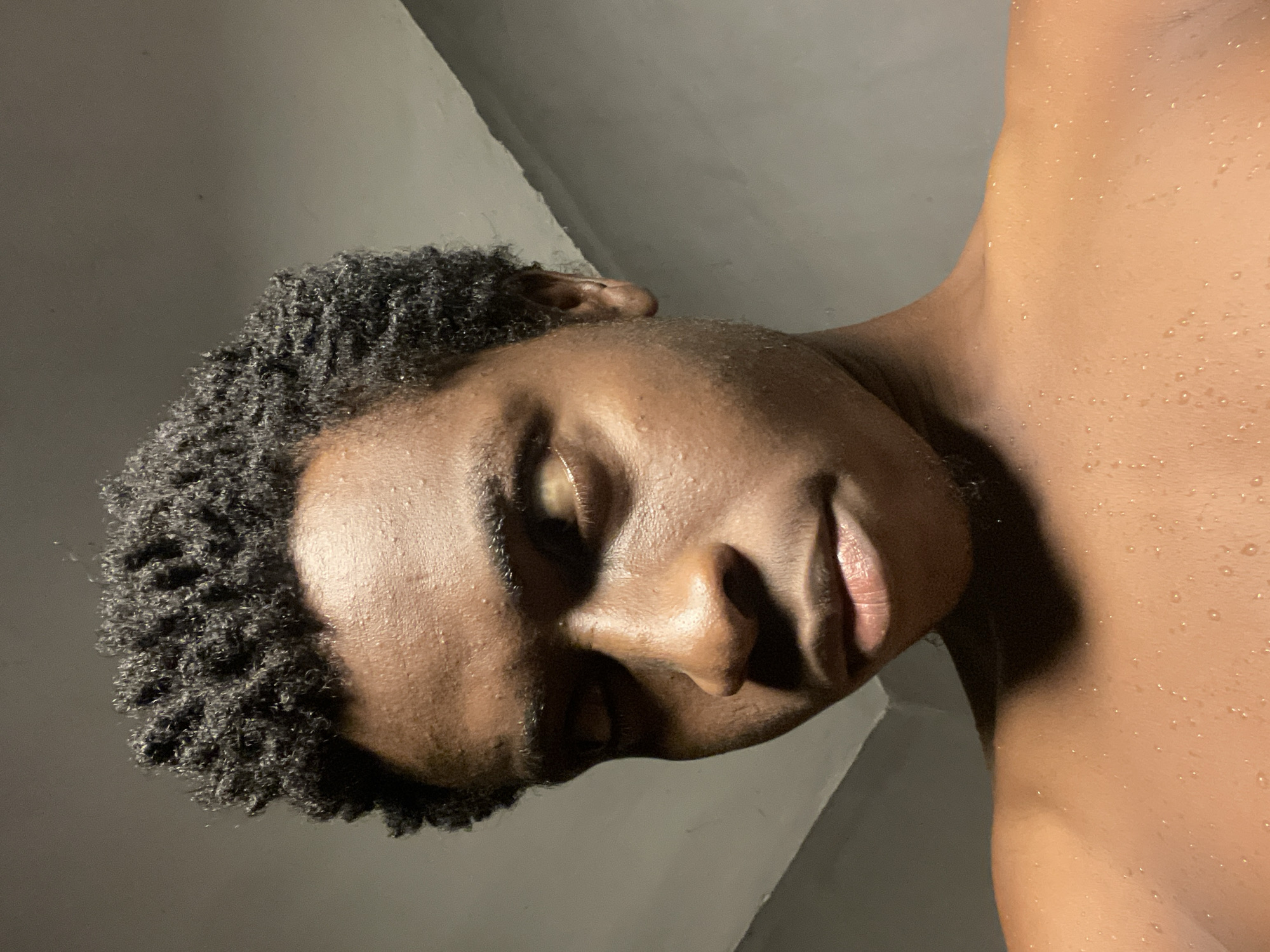 A close-up, low-angle shot captures a young Black male's face and upper torso in a dimly lit indoor environment. His skin is visibly damp, indicating perspiration or recent contact with water. He has short, dark, textured hair. His gaze is directed to the left, and his lips are slightly parted. The background consists of a plain, textured wall. The scene appears to be within a private residence in Nkpor, Nigeria, possibly a bathroom or bedroom.