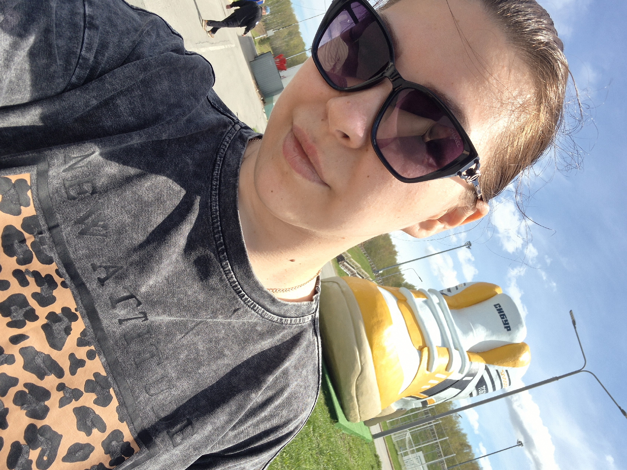 A woman wearing sunglasses and a t-shirt with a leopard print and grey top is taking a selfie. She is standing near a large sculpture of a boxing glove with the inscription "Сибур". In the background, there is a parking lot, a person walking, and a fence with trees behind it. The sky is blue with some clouds. This location is in the city of Tobolsk, Russia.