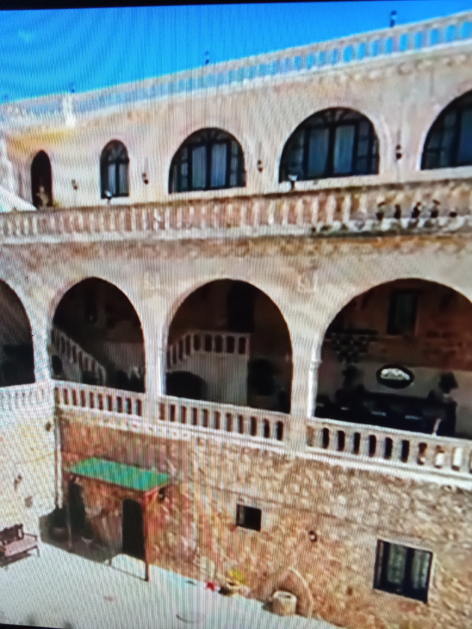 The image depicts a courtyard of a building with multiple arched openings and balconies. The building appears to be constructed of stone and has a light-colored facade. In the foreground, there is a covered area with a green roof and a dark opening, possibly an entrance. To the left of this structure, a bench or seating area is visible. The courtyard floor is paved. The architecture suggests a traditional or historic style. The surrounding environment is not clearly visible, but the overall impression is of an enclosed outdoor space within a building complex. The image's resolution is low, with visible pixelation and a grid-like pattern, indicating it might be a screenshot or a low-quality photograph. The scene is static, with no discernible people or activity. The question specifies the location as Рощин ауылдық округі, Kazakhstan, however, there are no visual cues within the image to confirm this location.