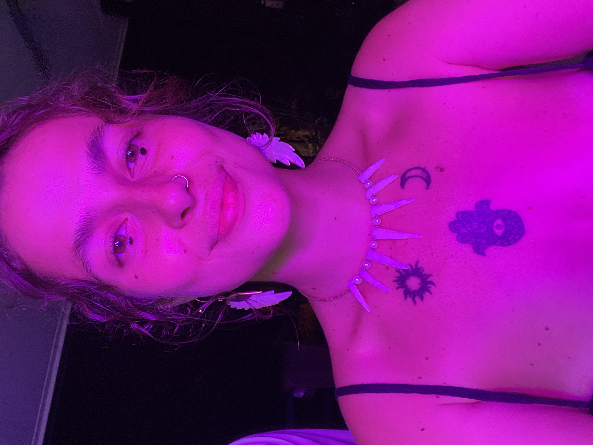 The image depicts a person with a nose ring and multiple ear piercings. They are wearing feather-shaped earrings and a necklace with moon, sun, and tooth-like charms. Two tattoos are visible on the upper chest, one a Hamsa hand and the other a sun. The lighting is predominantly magenta. There is no evidence to determine the location.