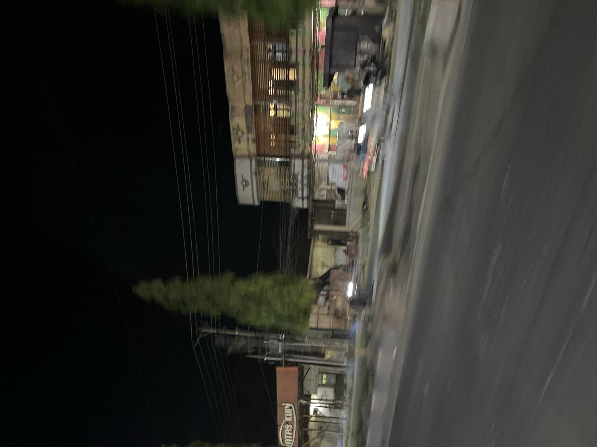 This is a blurry, low-light, handheld video clip taken at night, likely from a moving vehicle. The scene is a street in Bundar, Indonesia. The video is rotated 90 degrees clockwise, so the horizon appears vertical.
On the left side of the frame, there is a dark, open space, possibly a field or unlit area, with several power lines running vertically. To the right of the power lines, there is a row of buildings. These appear to be shops or businesses, with some windows illuminated from within. The architecture seems simple, with a mix of wooden and possibly brick facades. There are some signs and posters visible on the buildings, but they are too blurry to read clearly, except for one sign towards the bottom that appears to say "INTAS KUP". There are also trees or dense vegetation visible between the buildings and the power lines.
On the right side of the frame is a paved road, also blurry due to the motion of the vehicle. The road surface shows some wear and tear. The overall lighting is dim, with artificial lights from the buildings providing the main illumination. The blurriness suggests the video was captured quickly and without stabilization. There are no discernible people or specific activities taking place, other than passing by the scene. The time of day is clearly night, and the weather appears clear. The location cues are limited to the presence of shops, power lines, and a paved road, consistent with an urban or semi-urban street in Indonesia.