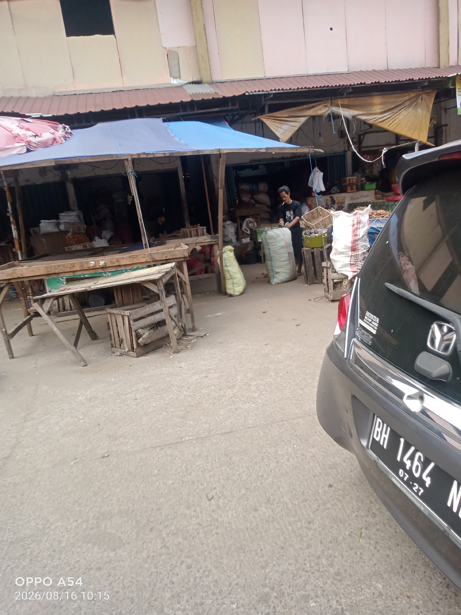 The image depicts an outdoor market scene in Jambi City, Indonesia. In the foreground, a black car's rear is visible, displaying a license plate with the characters "BH 1464 N". To the left of the car, a market stall with wooden structures and various goods is set up. A man wearing a dark t-shirt and pants is seen at the stall, holding what appears to be a cigarette. He is surrounded by sacks and baskets containing produce or other merchandise. Several other sacks are piled up behind the stall. A blue tarp covers part of the stall's structure, and a weathered brown tarp is draped over another section. The ground is paved with concrete. The background shows more market stalls and buildings, suggesting a bustling commercial area. The time stamp in the bottom left corner indicates "2026/08/16 10:15" and "OPPO A54".