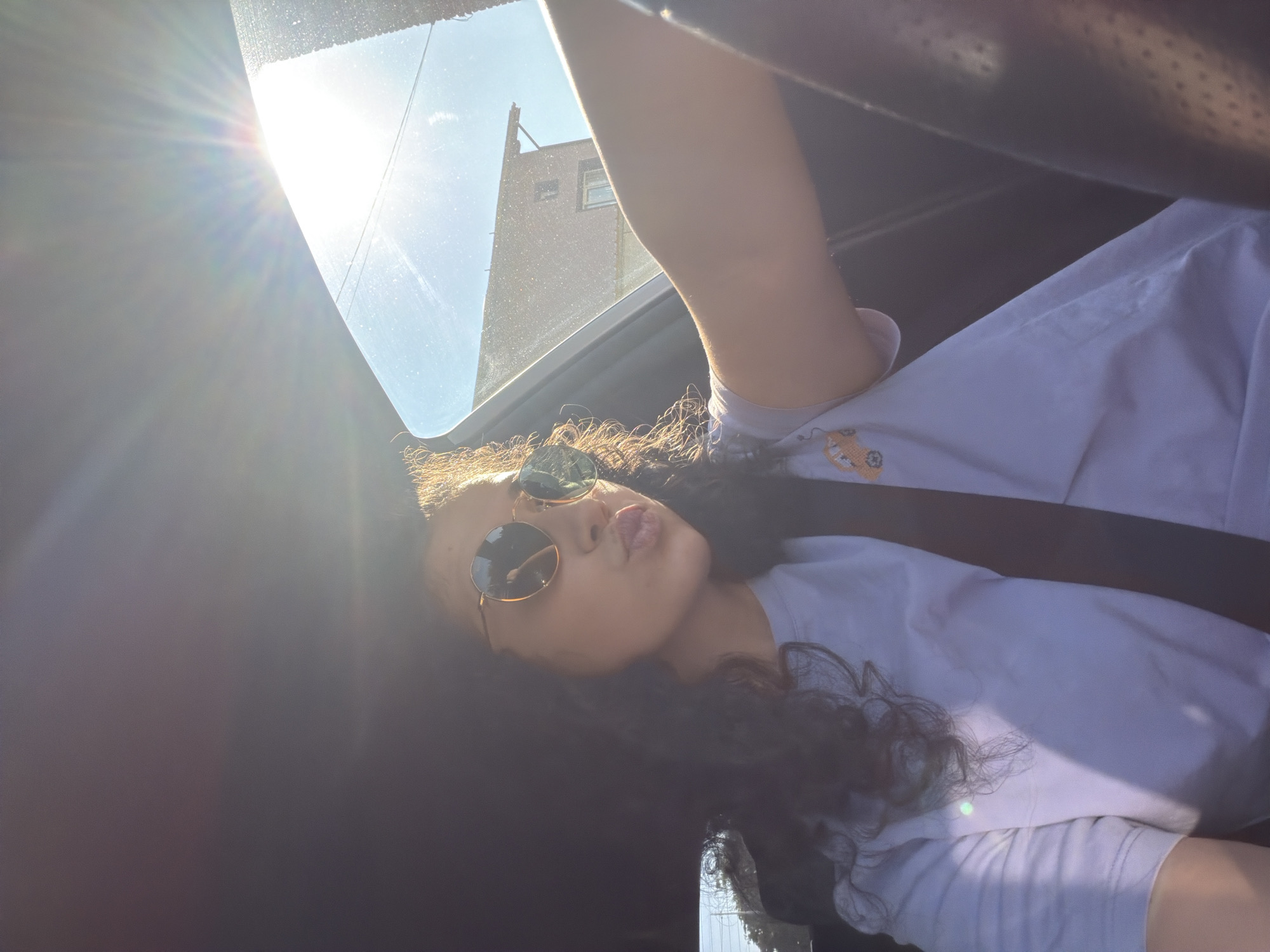 A young woman with dark curly hair and sunglasses is pouting her lips while sitting in the driver's seat of a car. The sun is shining brightly through the windshield, creating a lens flare. The woman is wearing a light purple t-shirt with a small orange car graphic on the left side. Her arm is extended, and she appears to be holding or reaching for something. In the background, out of focus, are buildings and a clear blue sky. The overall impression is a selfie taken while driving during the day in an urban or suburban environment.