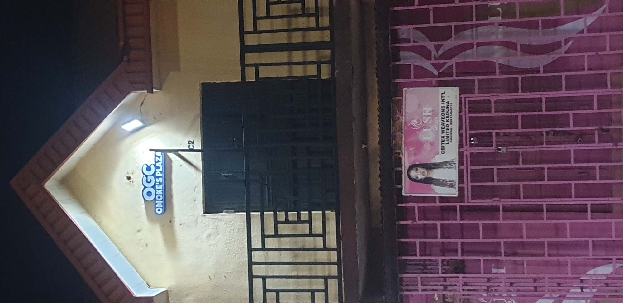 The image depicts a two-story commercial building at night in Narayi, Kaduna State, Nigeria. The upper level features a light-colored, possibly yellow, facade with a prominent gable roof. An illuminated blue sign on this level reads "OGC OMOKE'S PLAZA," with two white rectangular lights mounted on the facade above it providing illumination. Below the sign, a dark metal railing extends across the front of the upper floor. A dark pole or conduit is visible to the right of the sign, with "C2" inscribed near its base.
The lower level is covered by a closed, magenta-colored metal grate or gate. Attached to this gate is a white rectangular sign displaying the "LUSH Hair" logo, which features a woman's silhouette with flowing hair, next to an image of a woman with long dark hair. Below the logo, text reads "OBITEX WEAVEONS INT'L LIMITED KADUNA" and a partially visible phone number, "08033468512". A padlock is visible on the gate, and decorative white swirl patterns adorn the right side of the magenta gate.