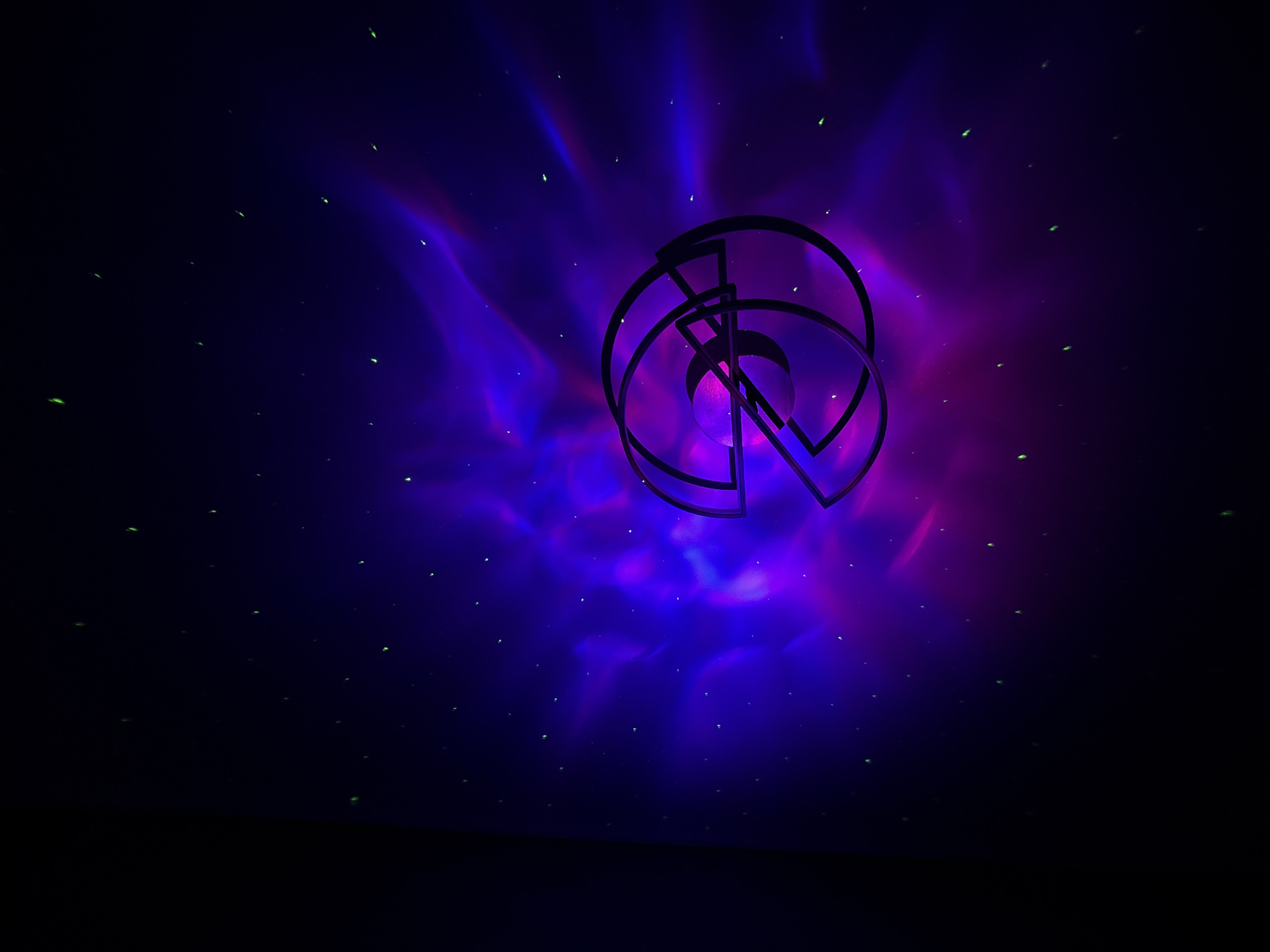This image depicts an abstract, dimly lit scene with a dark, amorphous foreground that could be a stage or a floor. Dominating the center is a stylized, metallic object, possibly a sculpture or a decorative element, composed of intersecting circular and linear forms. Behind this object, a vibrant nebula-like cloud of purple and pink light swirls, creating an otherworldly atmosphere. Scattered throughout the darker areas of the background are numerous small, bright green specks, resembling distant stars or particles. The overall mood is mysterious and slightly futuristic, with the interplay of dark shadows and luminous colors. There is no discernible activity or event taking place; the scene is static and atmospheric. No text is visible in the image. Given the location context of Chaikovsky, Russia, the setting could be an indoor venue or a specially designed space where this light and object installation is displayed. The lighting suggests a nighttime or controlled indoor environment.