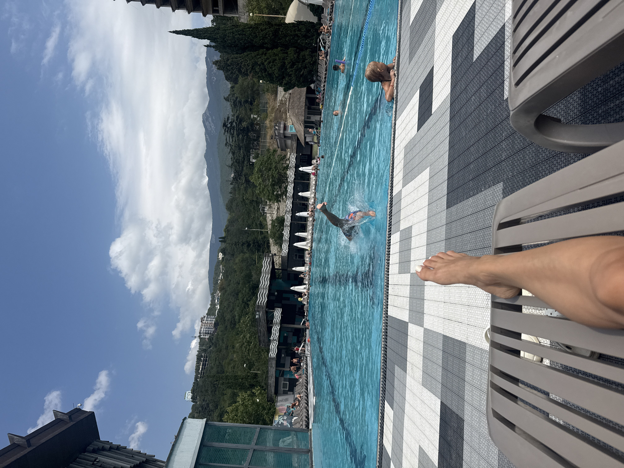 The image depicts a swimming pool area at a resort, likely in Yalta, Ukraine, given the mountainous backdrop. In the foreground, a person's legs and feet are visible, resting on a lounge chair. The pool is a vibrant turquoise blue, with several lanes marked by ropes. A person is captured mid-air, diving into the pool, with water splashing around them. Other individuals are visible in the pool, some swimming and others relaxing. The poolside is lined with lounge chairs, some occupied. In the background, there is a building with a dark, angled roof, and beyond that, lush green hills and mountains under a partly cloudy sky. The overall scene conveys a leisurely summer atmosphere.