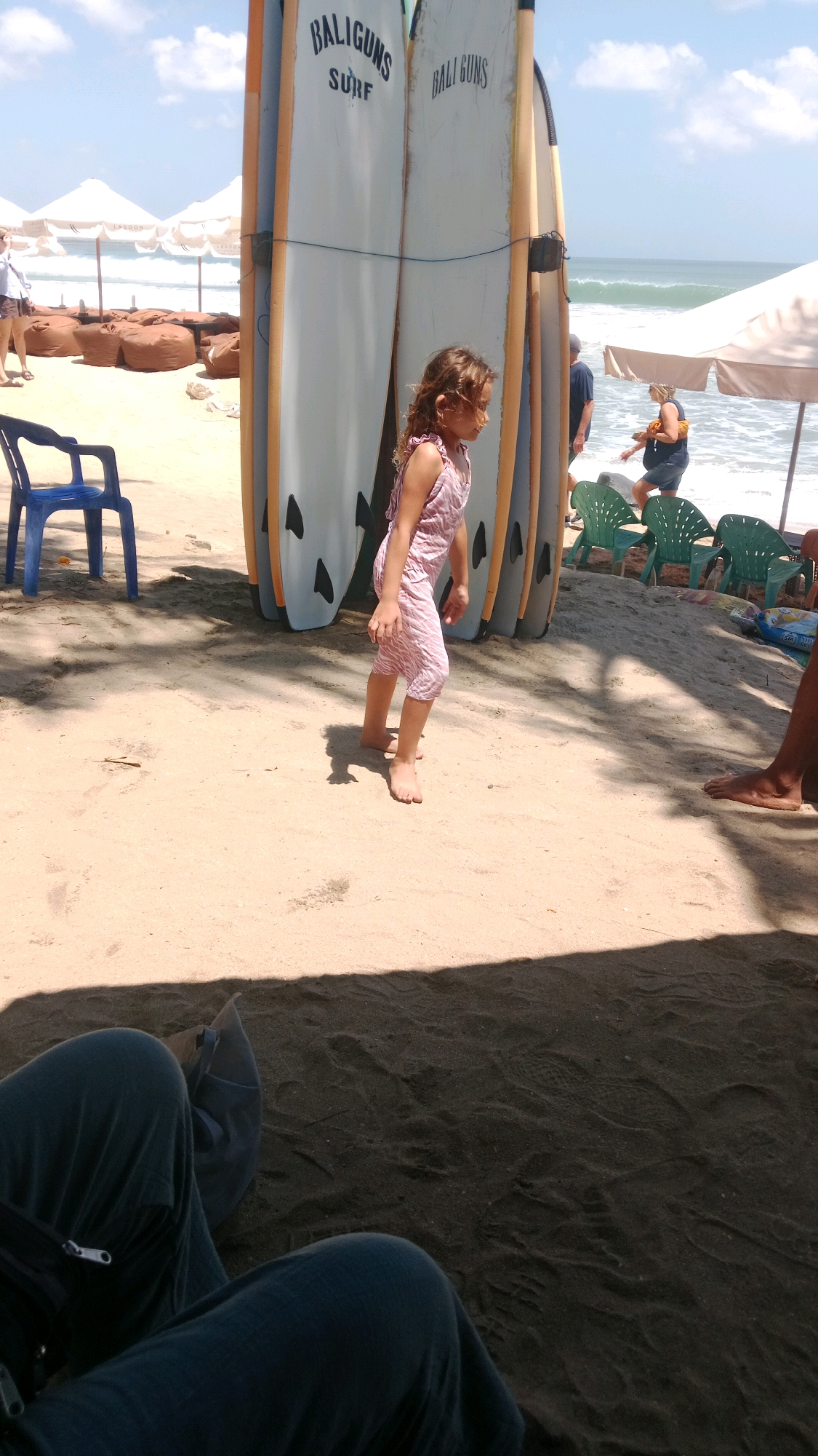 A young girl in a pink, patterned romper walks barefoot on a sandy beach in Kuta, Indonesia. She is positioned to the right of two tall surfboards with "BALIGUNS SURF" and "BALI GUNS" written on them. Behind the surfboards, people are visible by the ocean, with waves rolling in under a bright, partly cloudy sky. Beach umbrellas and chairs dot the coastline. In the foreground, the legs and lap of someone seated on the sand are visible, casting a shadow. The scene suggests a warm, sunny day, likely in the afternoon, with a relaxed beach atmosphere.